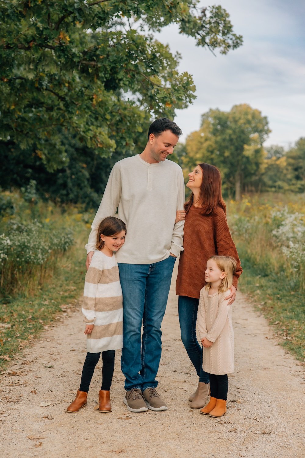 A family of five enjoying an outdoor walk on a dirt path surrounded by trees and greenery, smiling and looking at each other.