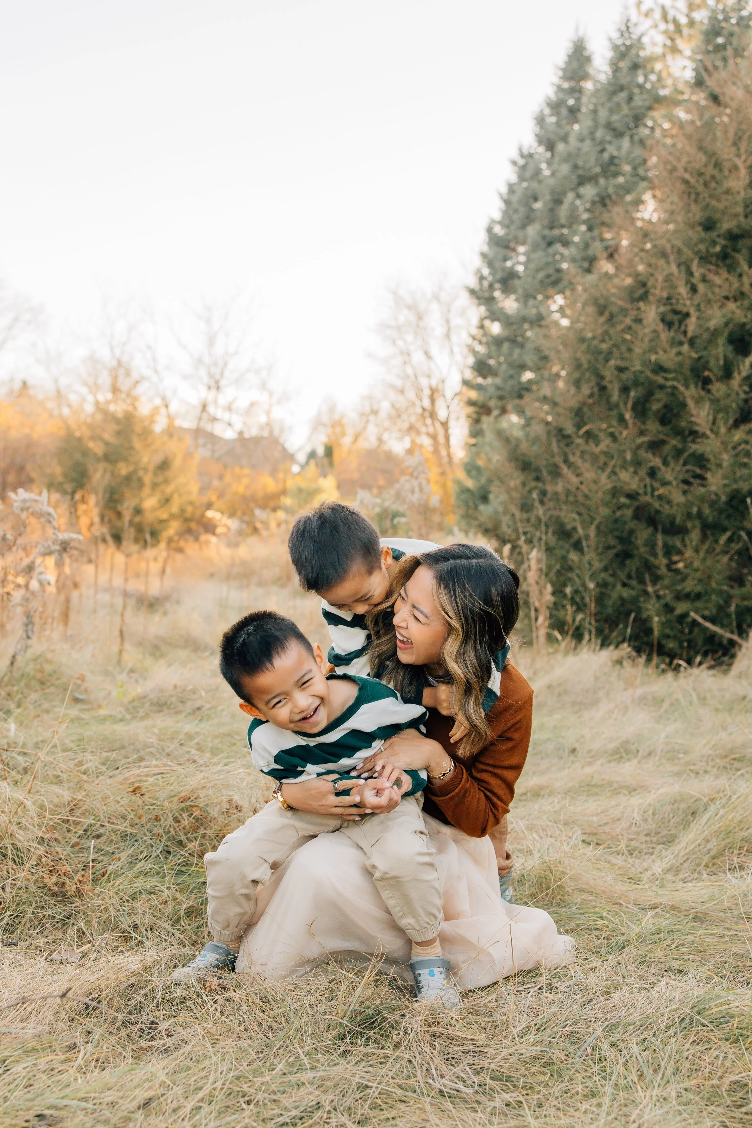 A woman and two young boys playing and laughing in a grassy field with trees in the background during autumn.