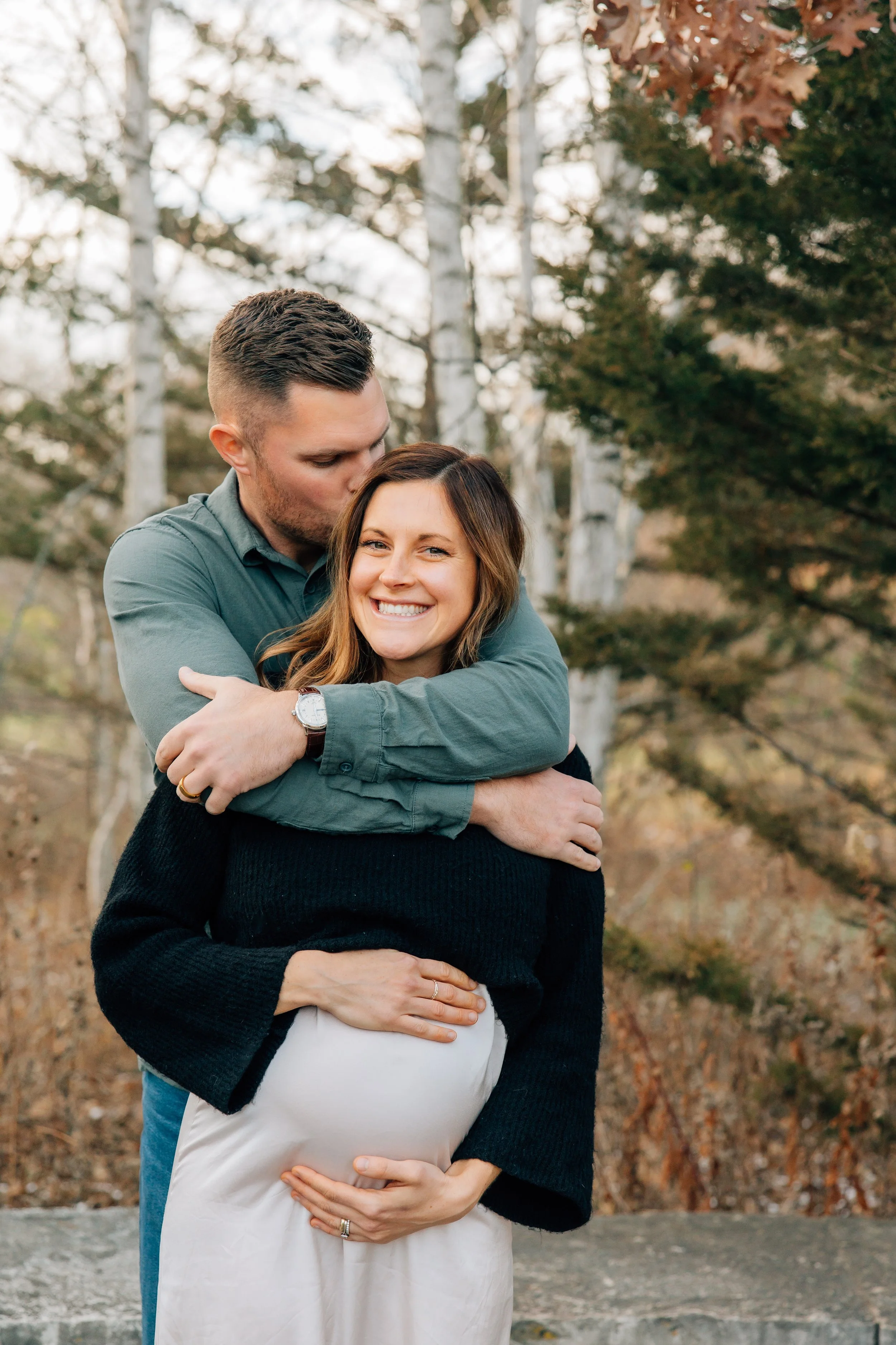 A happy pregnant woman smiling while a man kisses her on the head, outdoors among trees in fall weather.