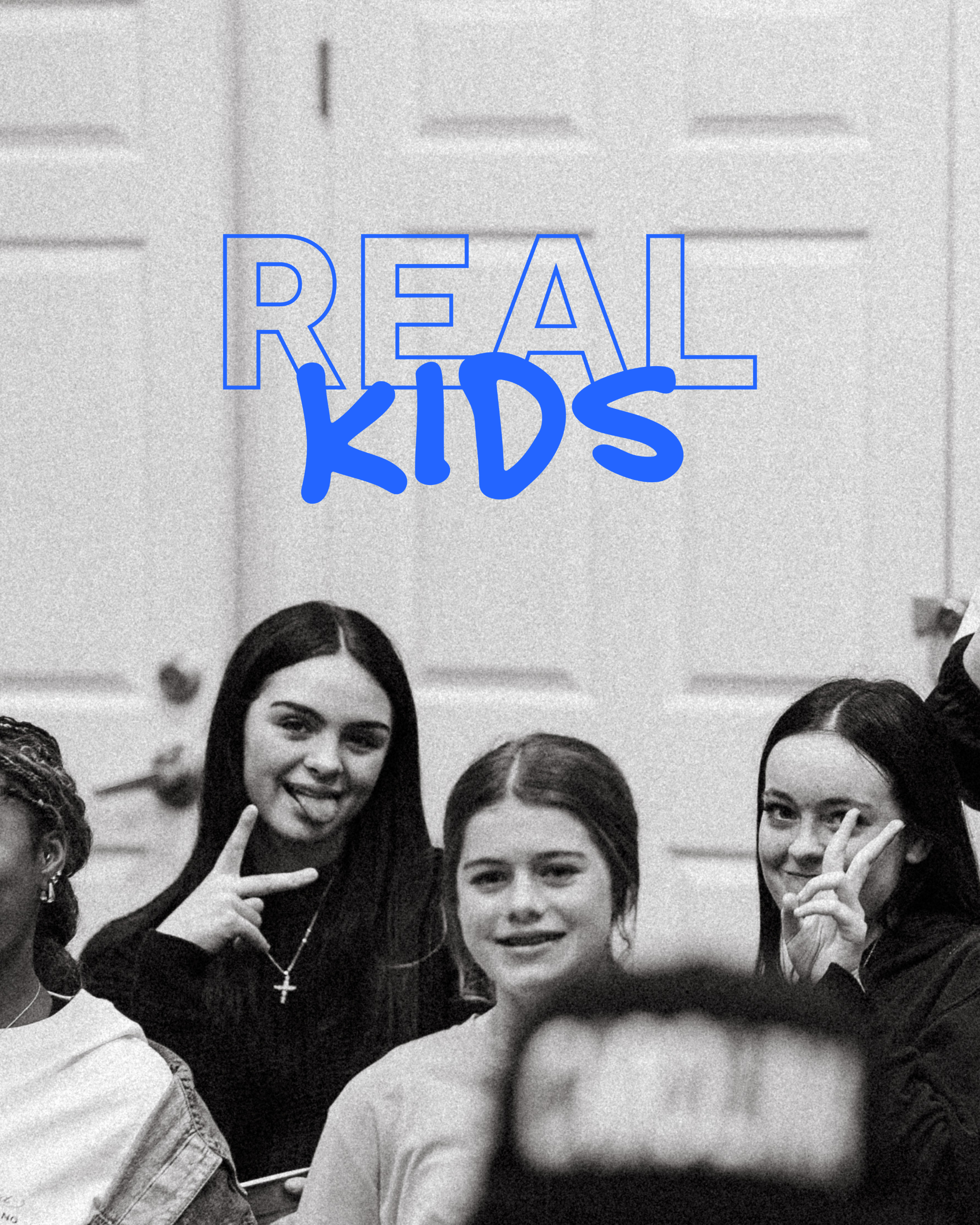 Black and white photo of several girls, one in the center forming a peace sign with her fingers, wearing a blazer and cross necklace, against a background with a large sign that reads 'REAL KIDS' in blue outline.