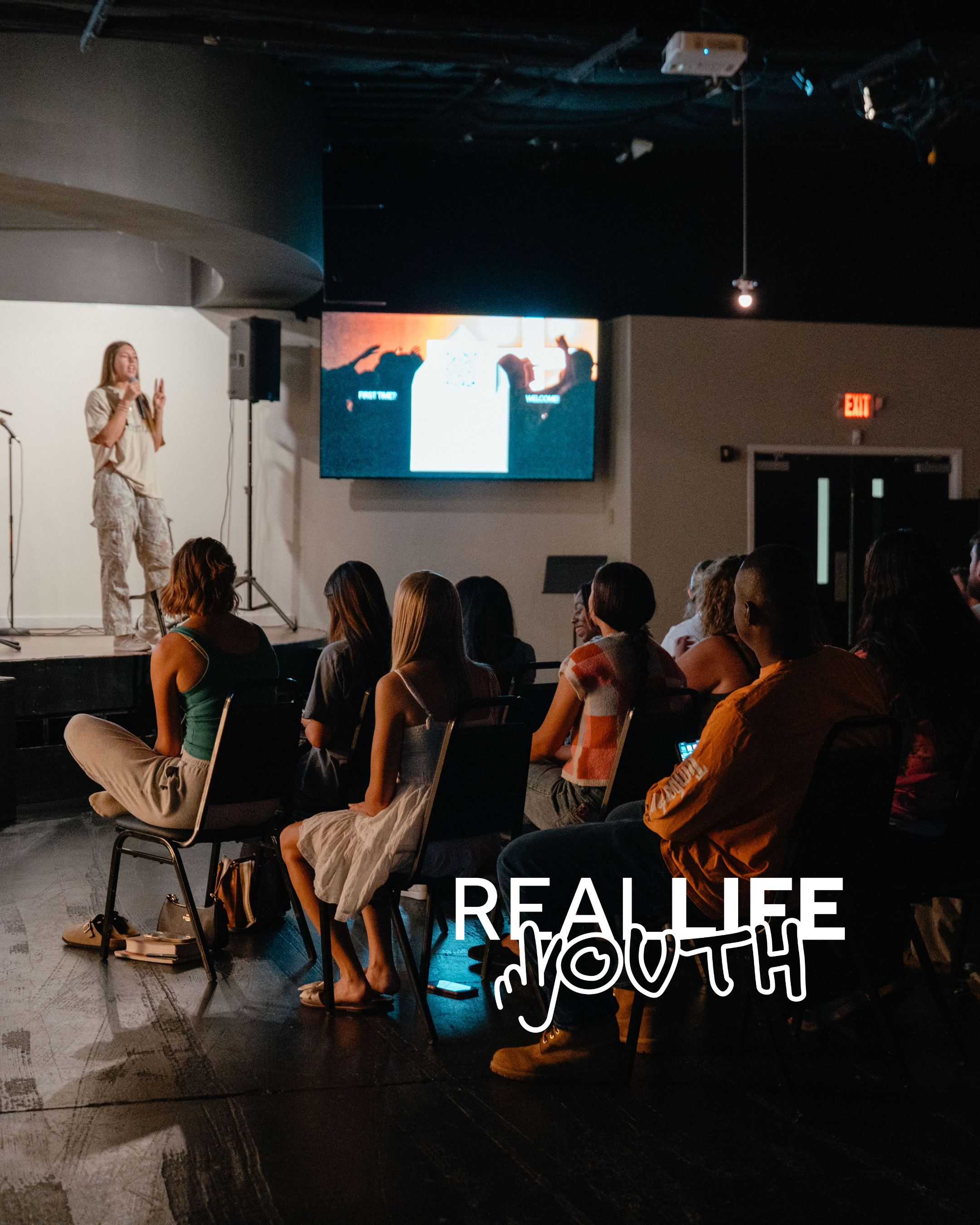 A woman is speaking on stage with a microphone, wearing beige camouflage pants and a beige shirt. She is gesturing with her hands, showing two fingers. There is a large screen behind her displaying an image with a silhouette of people taking photos and the text 'First Time?' and 'Welcome.' The audience is seated, some with crossed legs, in a dark room with a projection system hanging from the ceiling and an emergency exit sign on the wall.