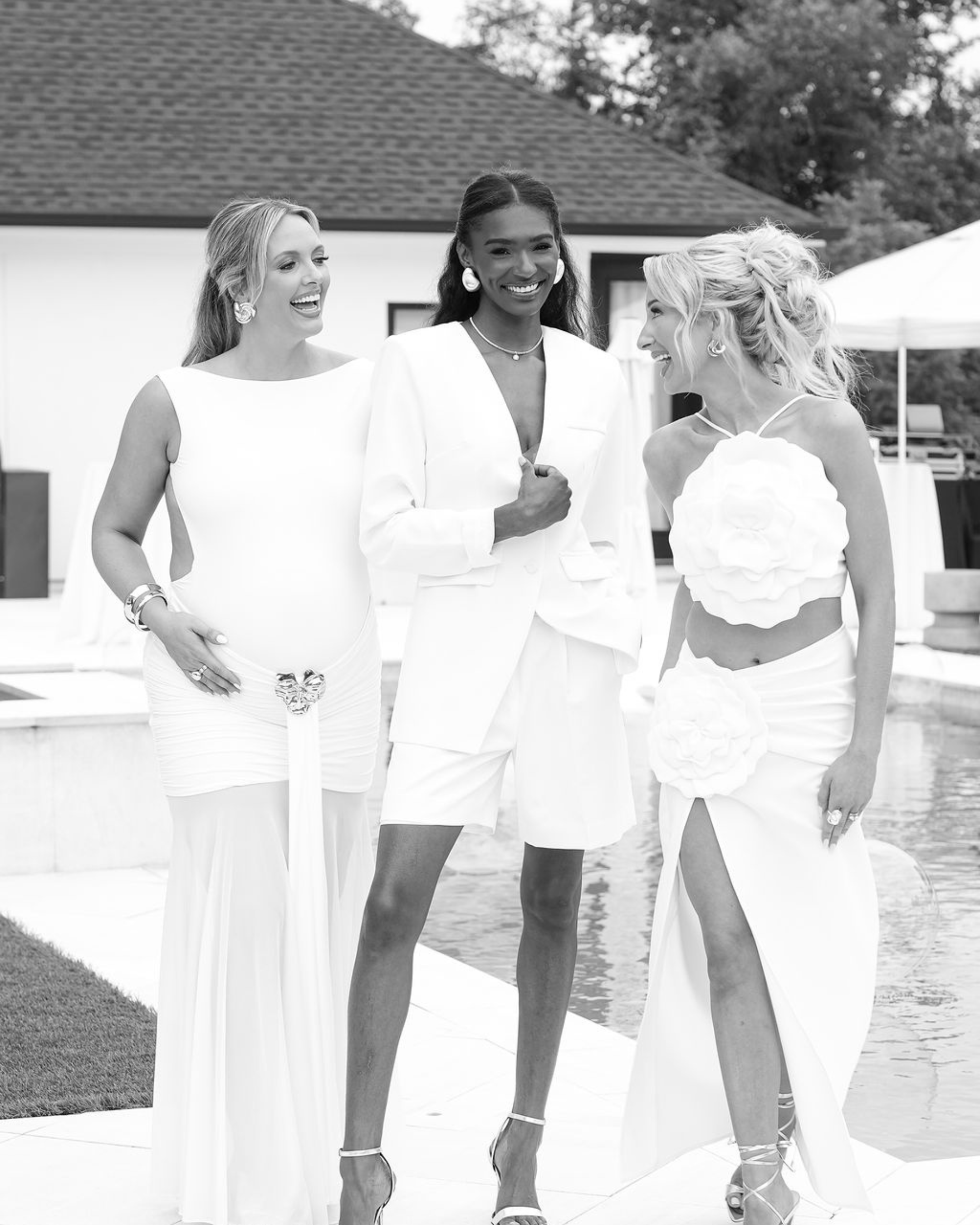 Four women smiling and laughing outdoors near a swimming pool, dressed in elegant white outfits and high heels.