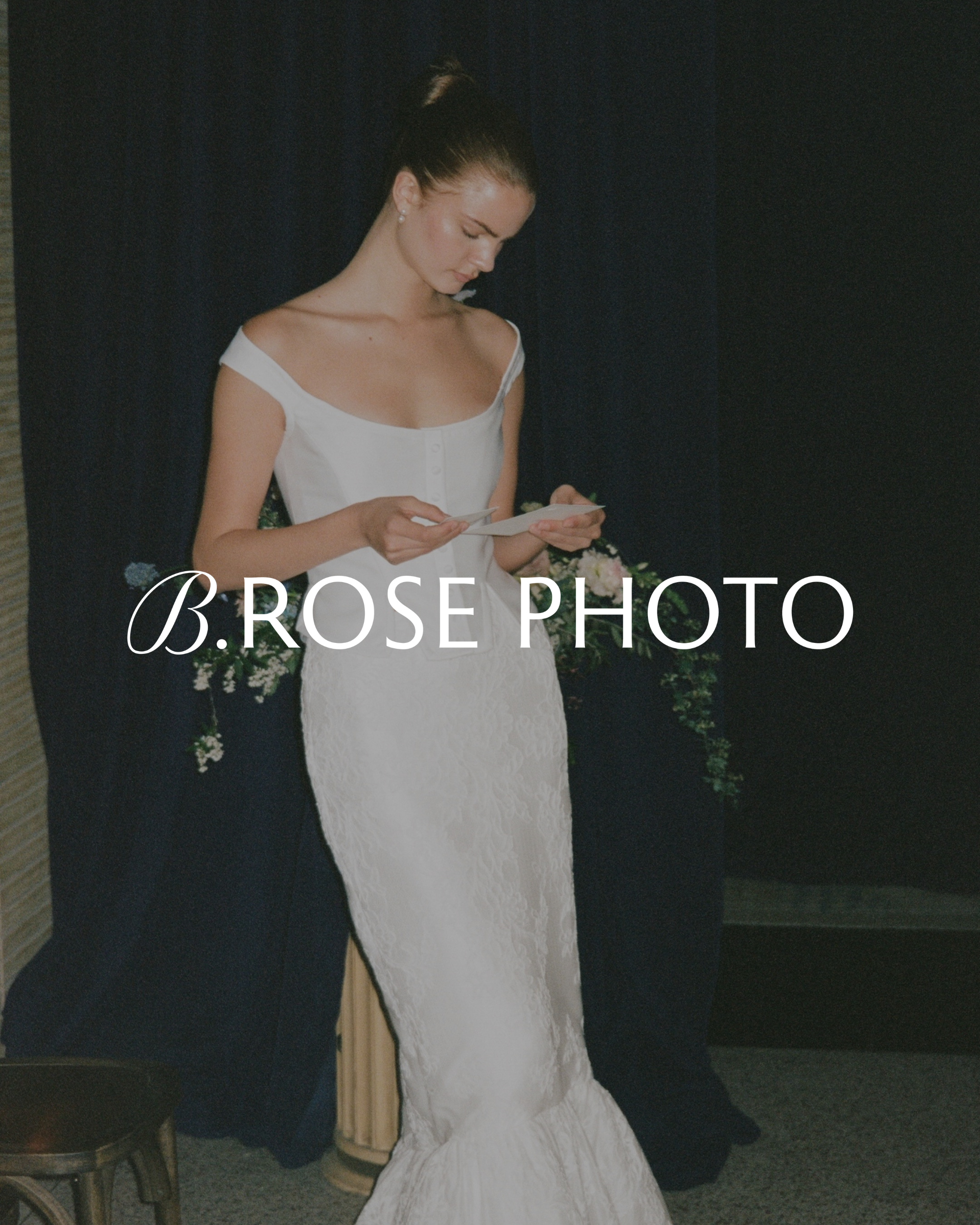 A woman with dark hair tied up in a bun, wearing pearl earrings, in a white dress, holding a piece of paper, against a dark background with floral arrangements.