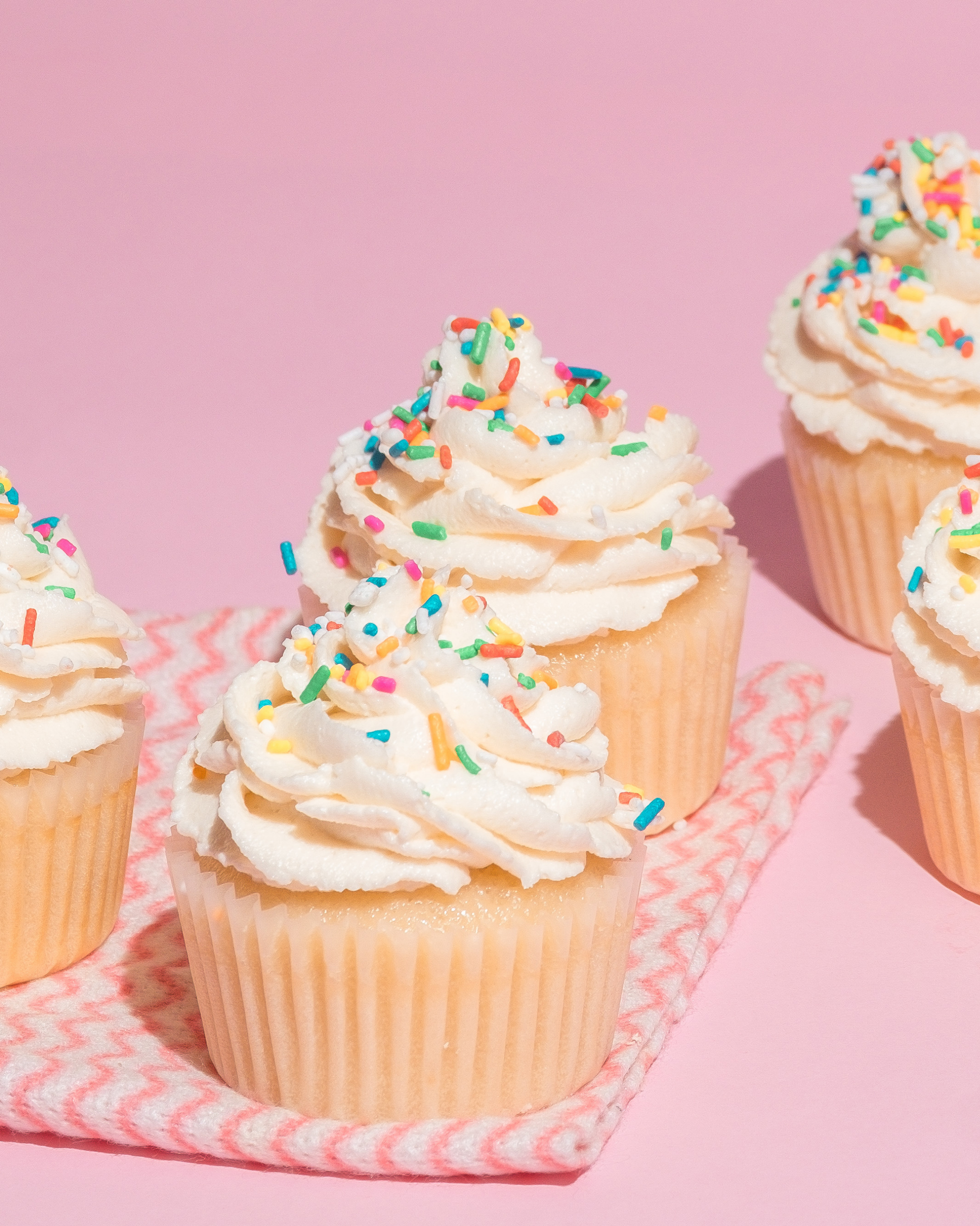 Close-up of three cupcakes with white frosting and colorful sprinkles on a pink background, placed on a pink and white striped cloth.