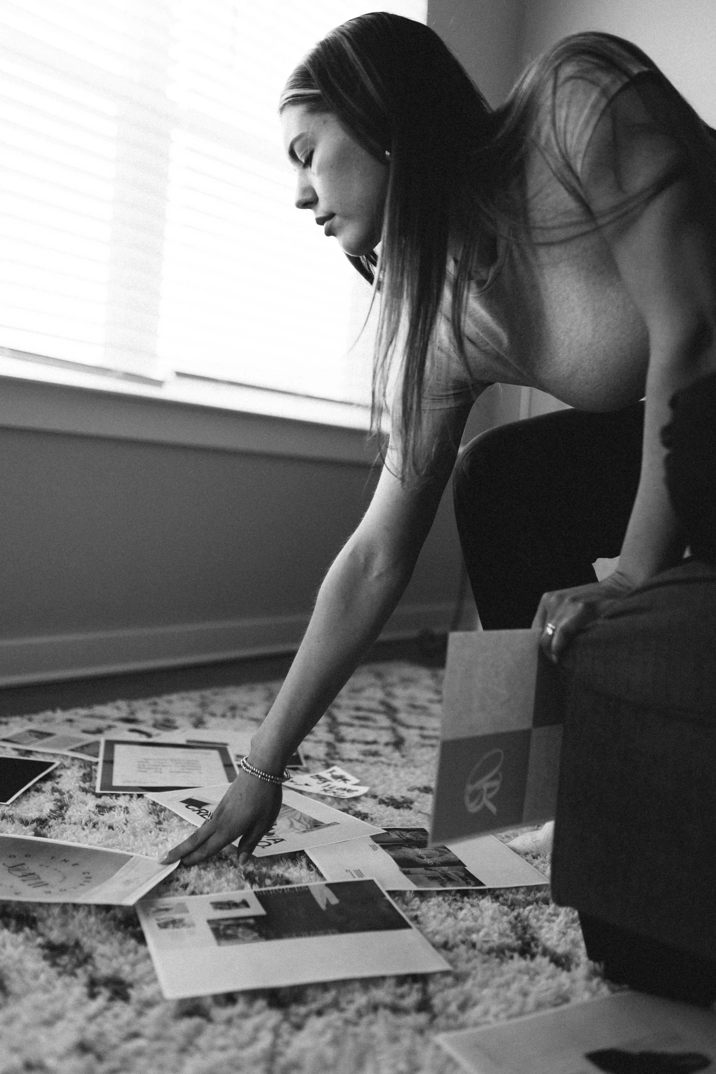 A woman with long hair leaning forward, reaching out to various papers and magazines spread on a carpeted floor, with a window with blinds in the background.