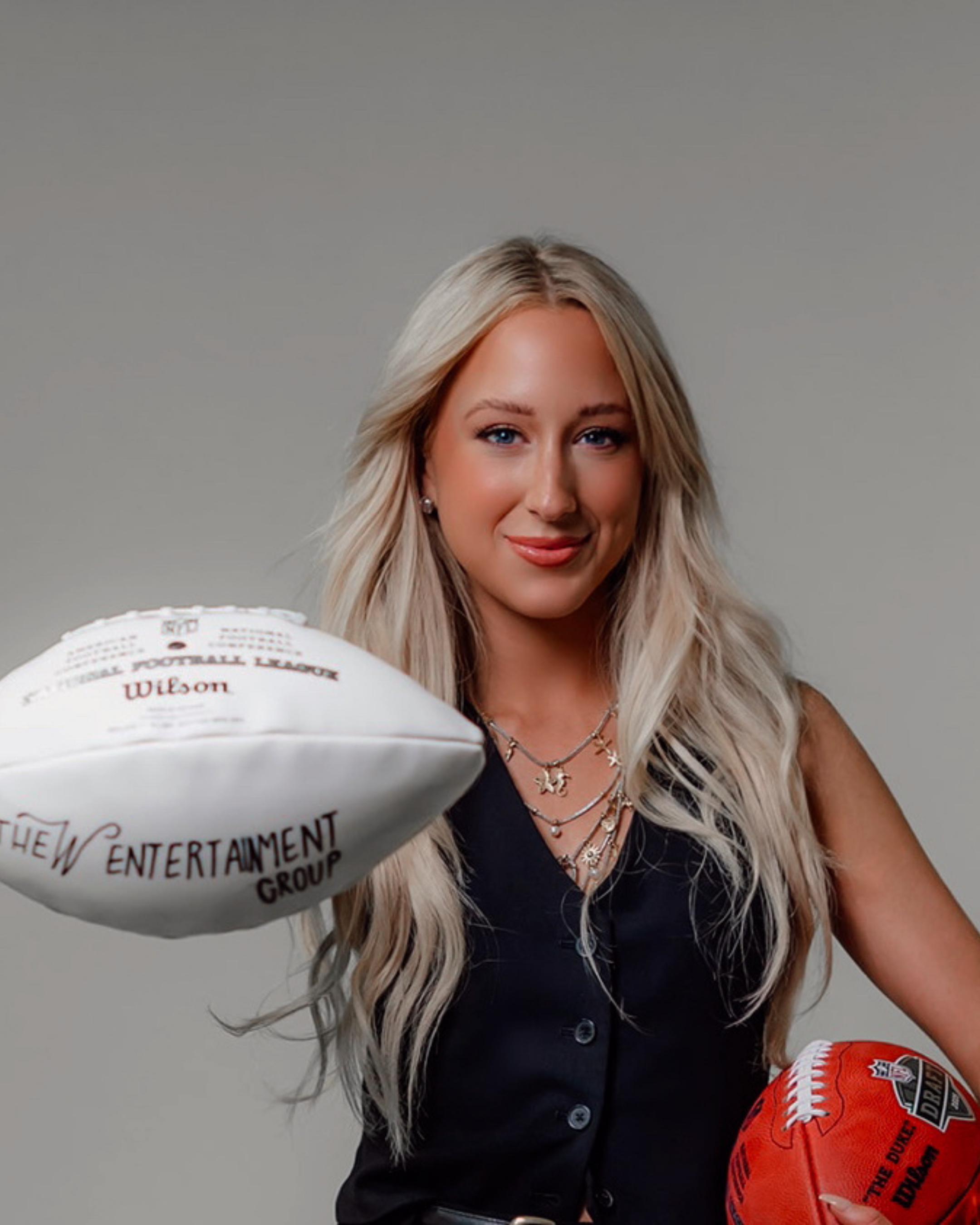 A smiling woman with long blonde hair holding an orange football with logos and text. She is wearing layered necklaces and a black sleeveless top, standing against a neutral gray background.