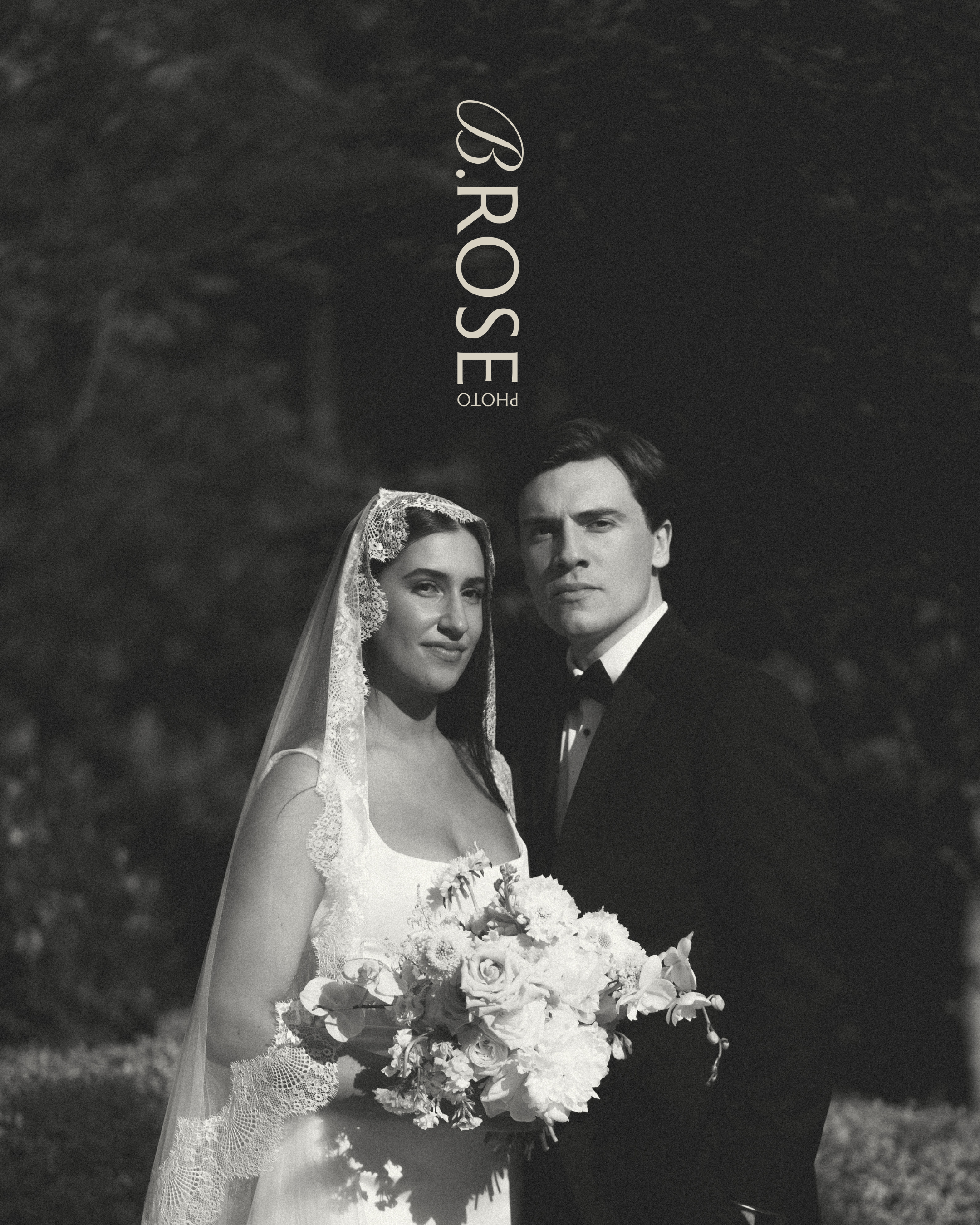 A black and white photo of a bride and groom in wedding attire, standing outdoors. The bride is wearing a lacy wedding dress and a veil, holding a bouquet of flowers. The groom is wearing a tuxedo. They are looking at the camera with serious expressions.