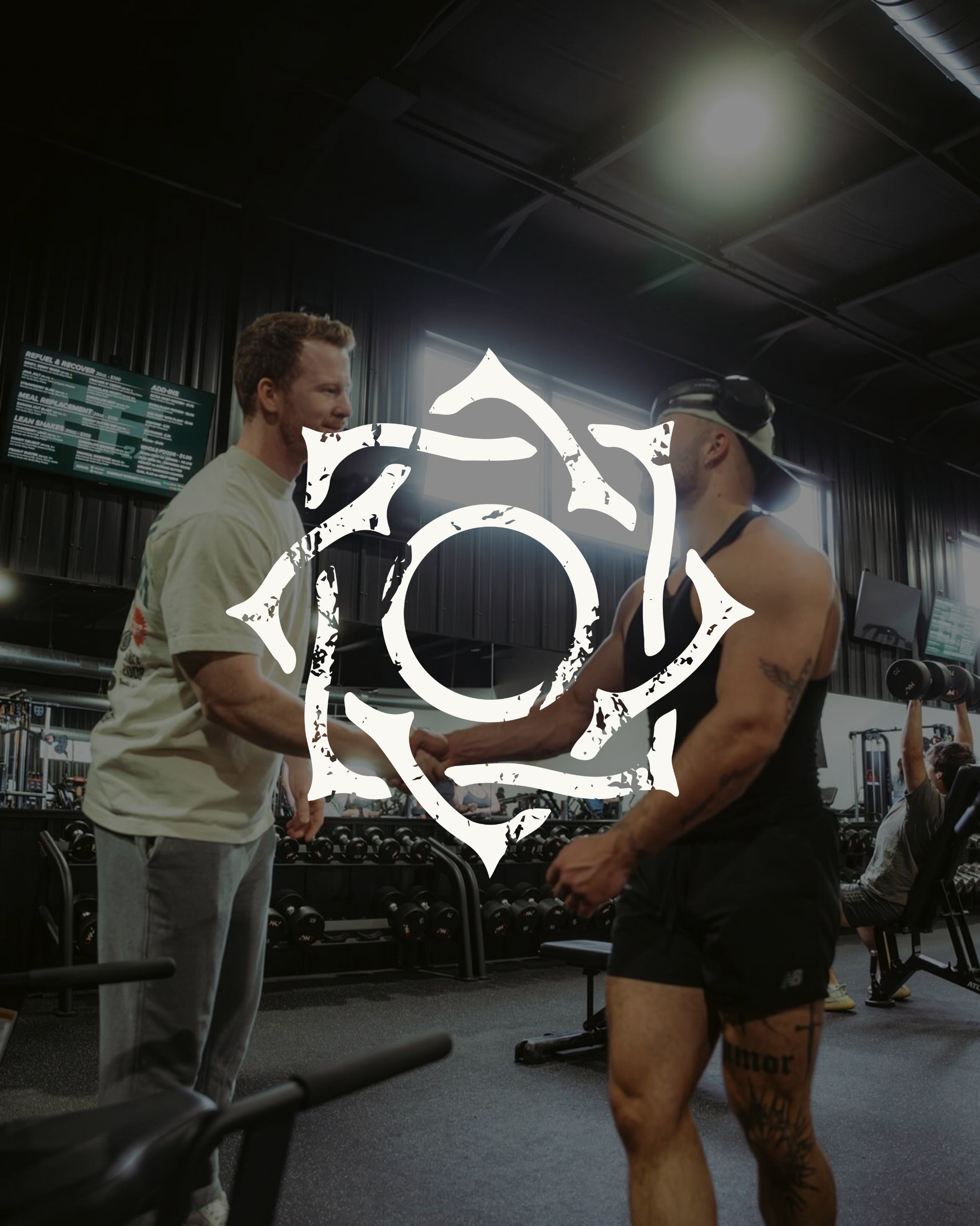 Two men shaking hands in a gym, one is shirtless with tattoos and the other is wearing a white t-shirt, with a wall of dumbbells and a menu board behind them.