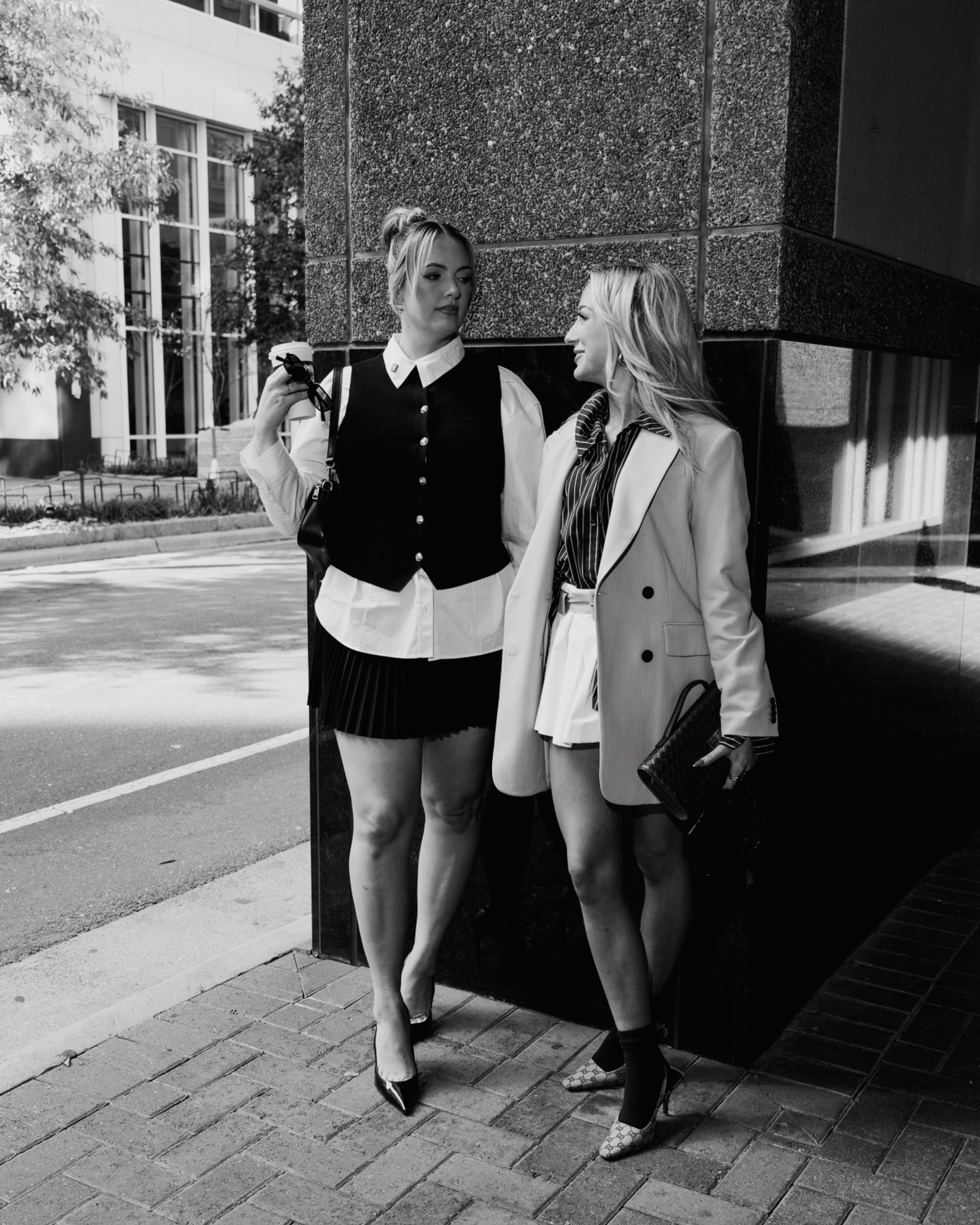 Two women dressed in professional attire are standing on a city sidewalk beside a large building. They are engaged in conversation and leaned against a reflective, dark-colored pillar. One woman is holding sunglasses and a purse, and both are wearing high-heeled shoes and stylish outfits.