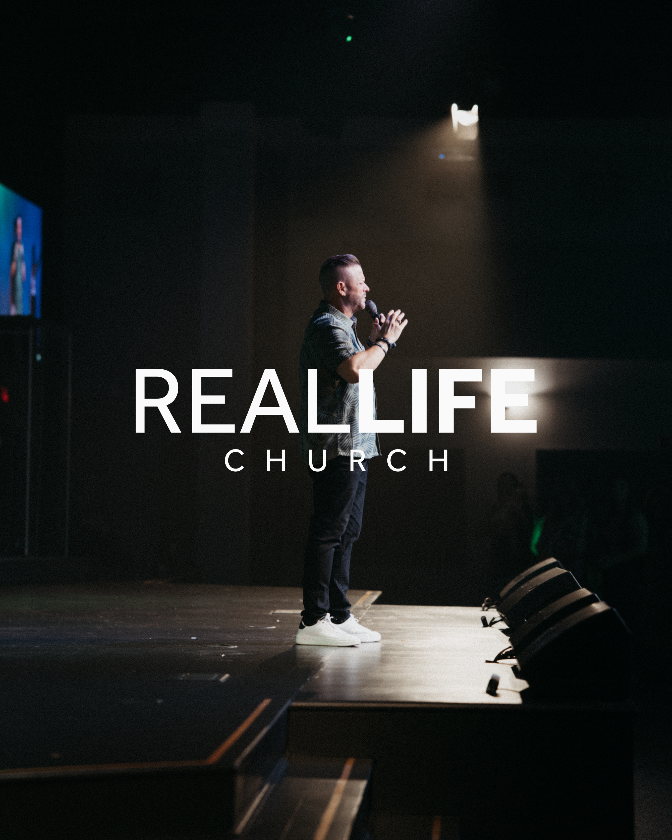 A man speaking into a microphone on stage at a church event, with the words 'REAL LIFE' and 'CHURCH' visible in the background.