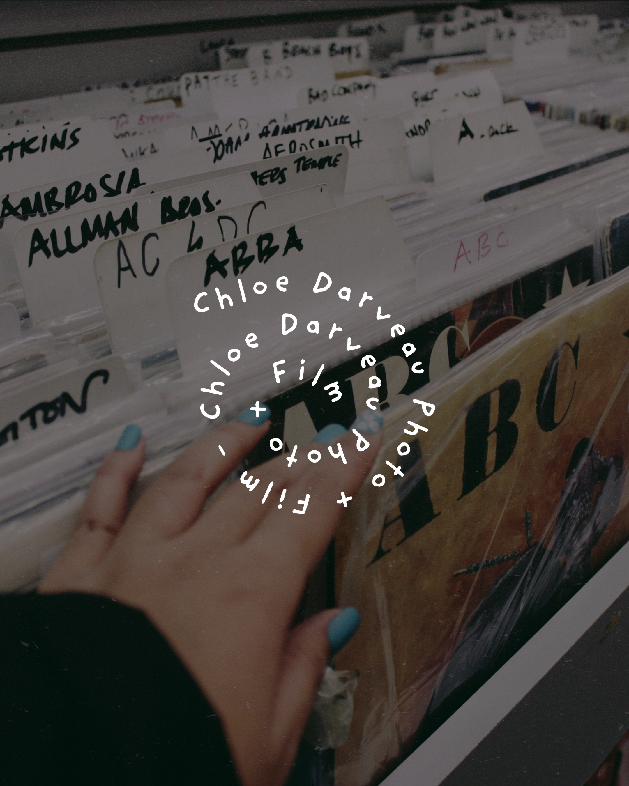 A person's hand with blue nail polish is pointing at a row of vinyl record sleeves in a store. The record sleeves have handwritten labels indicating their content, such as 'AMBROSIA' and 'ABC'.