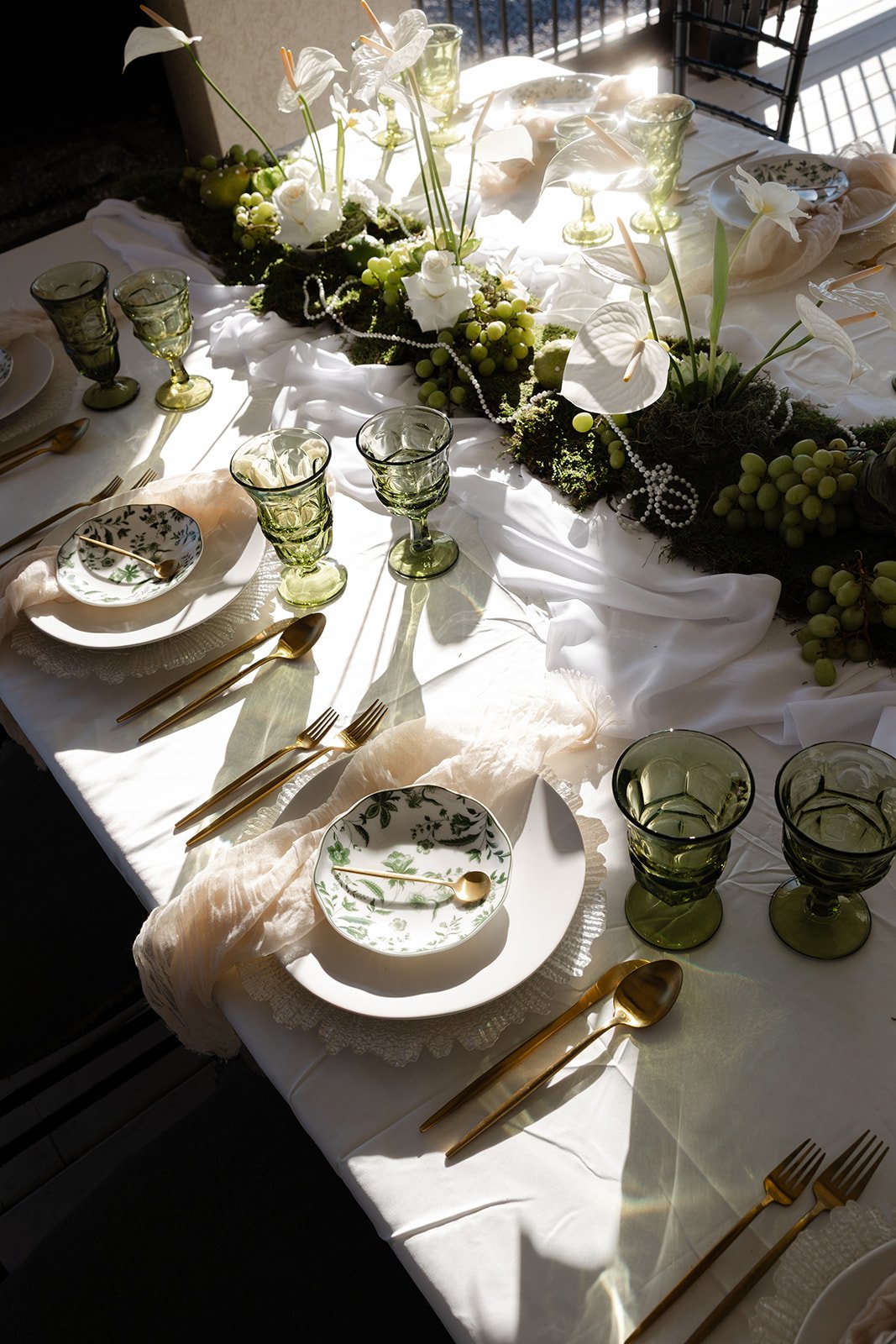 Elegant dining table decorated with a white tablecloth, gold flatware, green wine glasses, white plates with green floral patterns, and a floral centerpiece with white flowers, green grapes, and greenery.