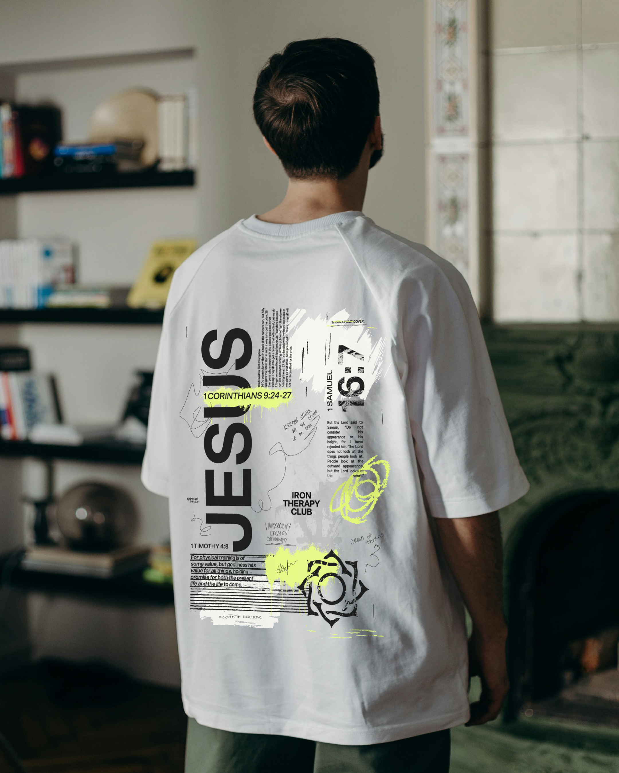 The image shows a man from the back, wearing a white t-shirt with text and graphics, standing indoors near a window with decorative tiles visible on the wall. Shelves with books and other items are in the background.