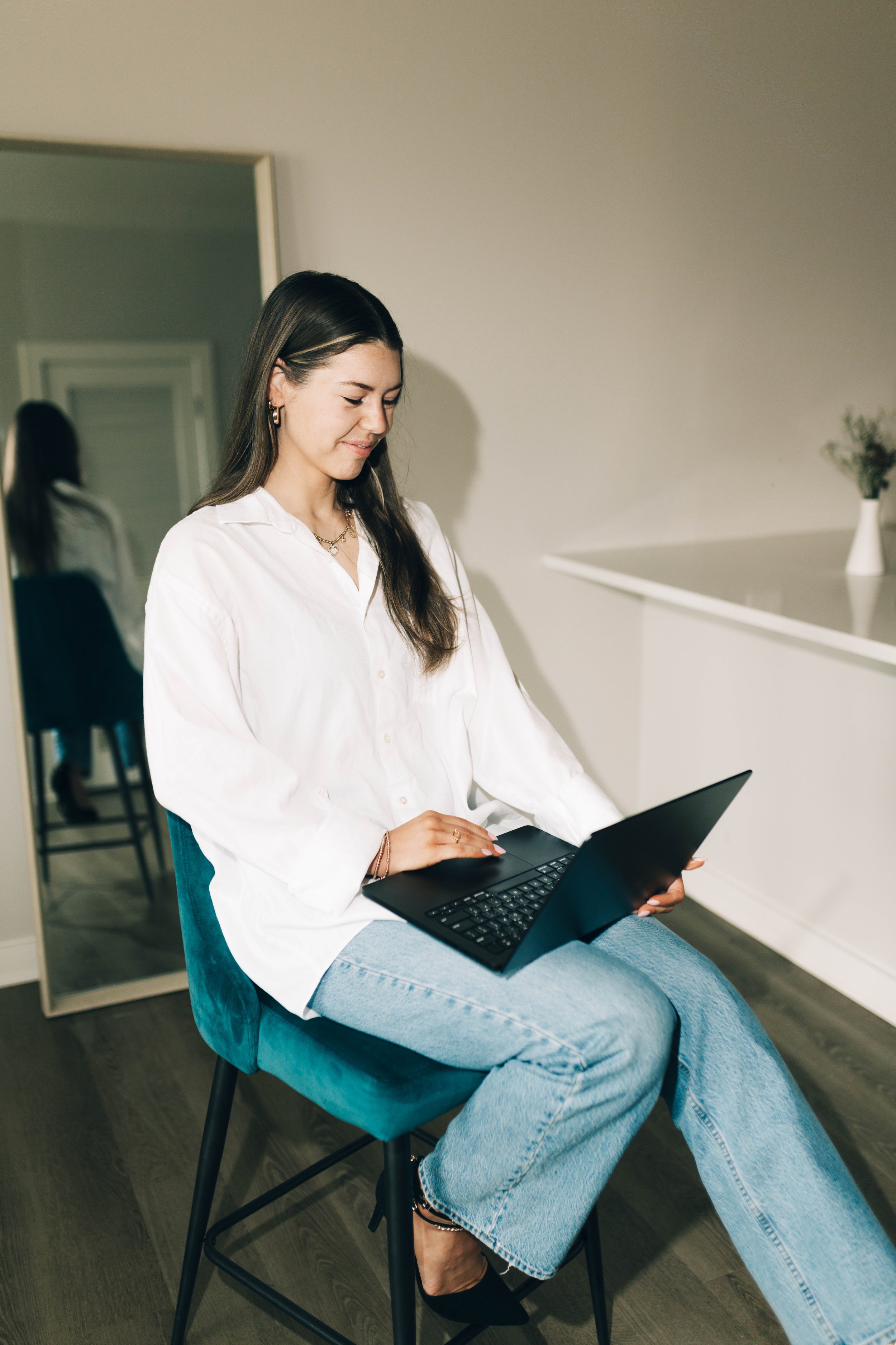 A young woman with long brown hair, wearing a white shirt and light blue jeans, sitting on a teal chair, working on a black laptop inside a modern, minimalistic room.