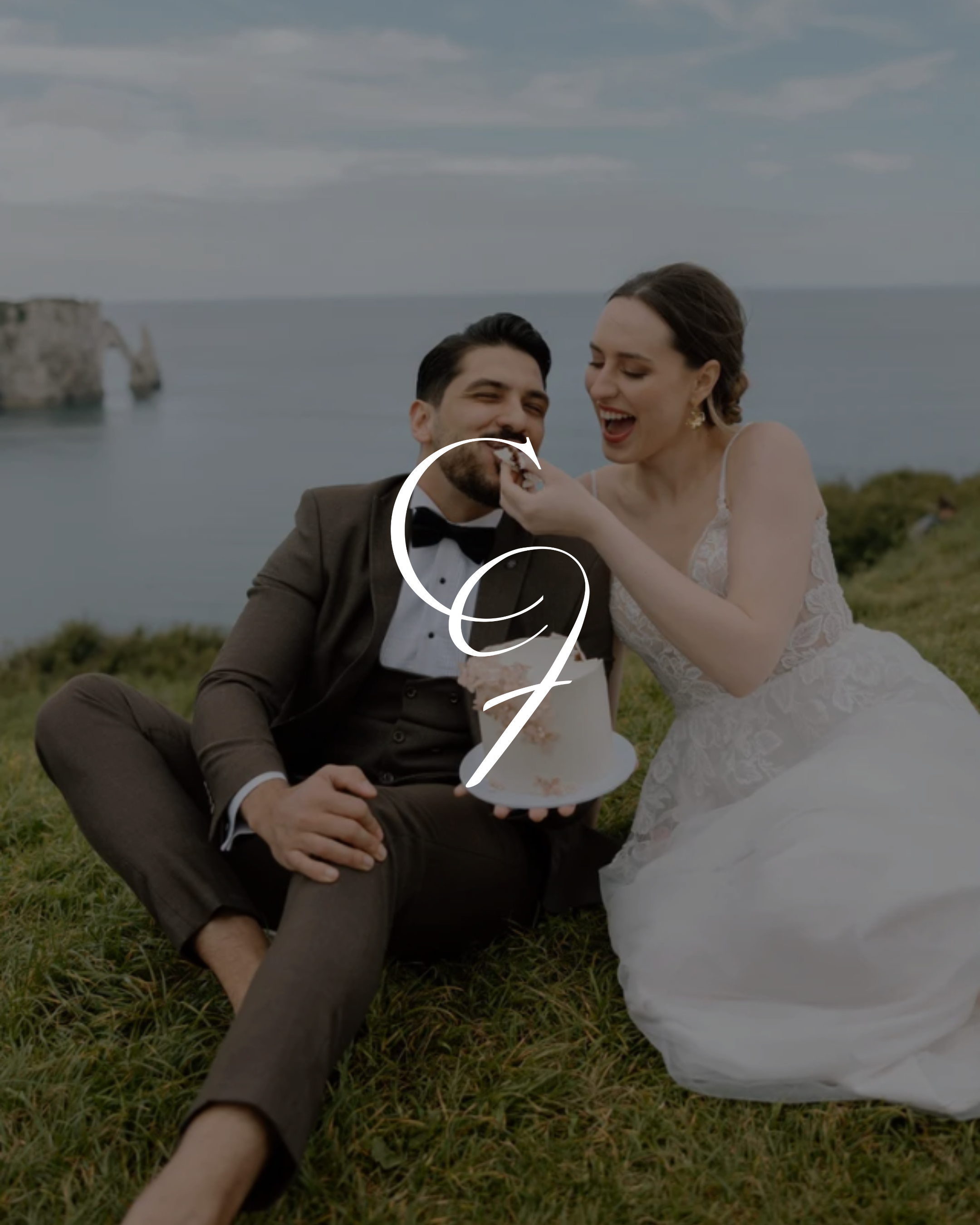 A newlywed couple with light skin and dark hair sitting on grass outdoors by the sea; the bride in a lace wedding dress and the groom in a tuxedo, sharing a piece of cake.