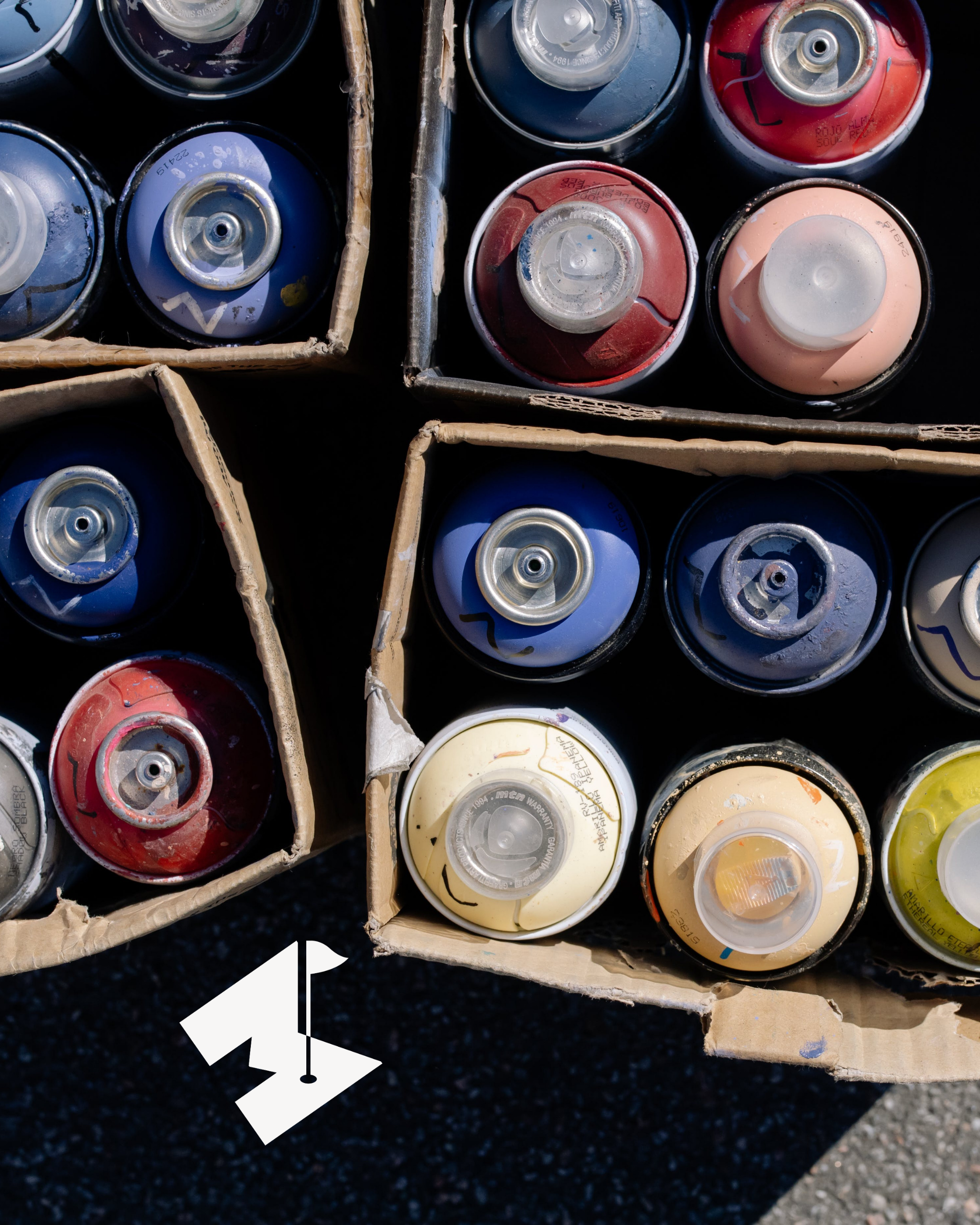 A close-up of a cardboard box filled with spray paint cans, organized by color, with a focus on blue, red, white, yellow, and beige cans.