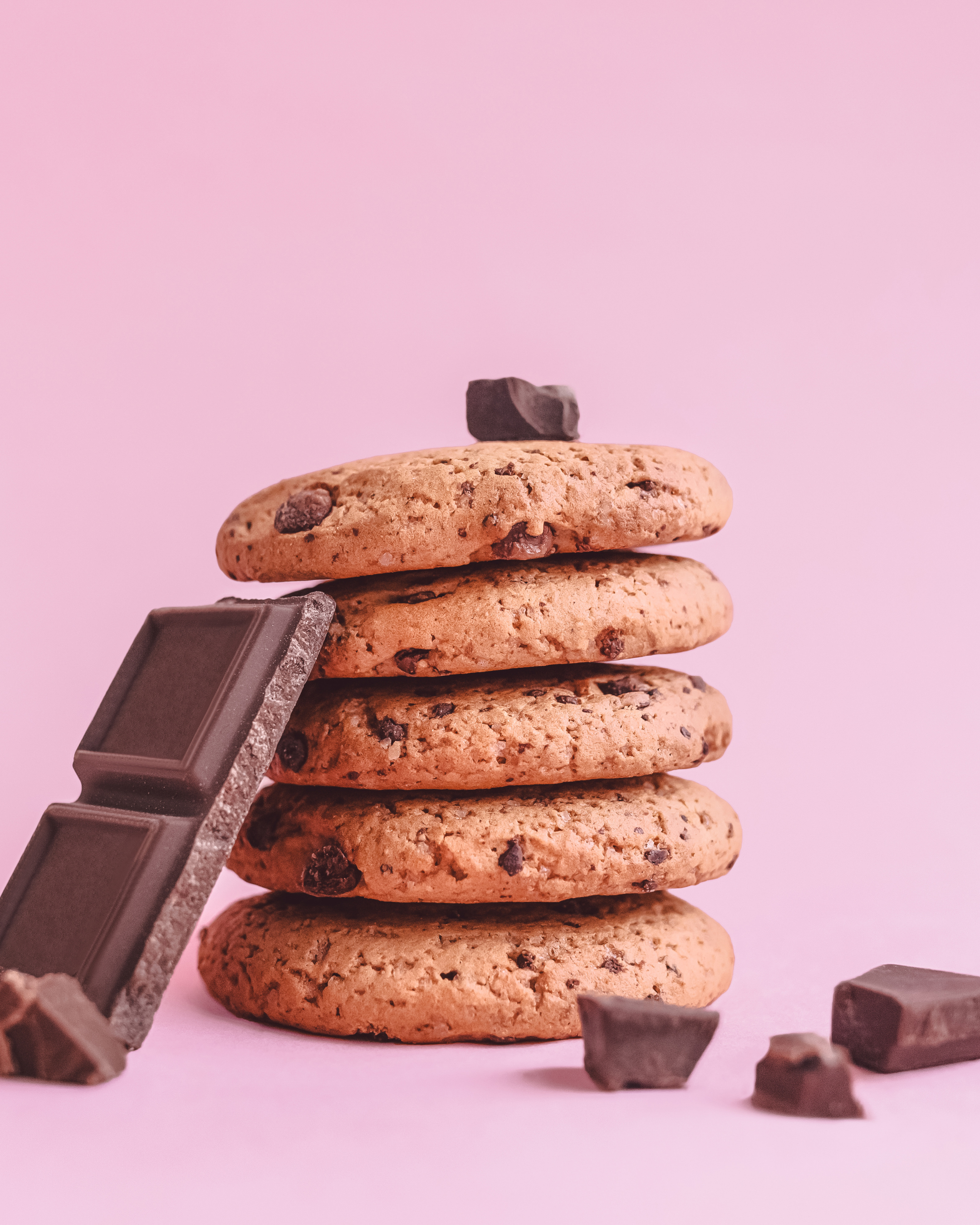 Stack of four chocolate chip cookies with broken pieces of chocolate in the foreground against a pink background.