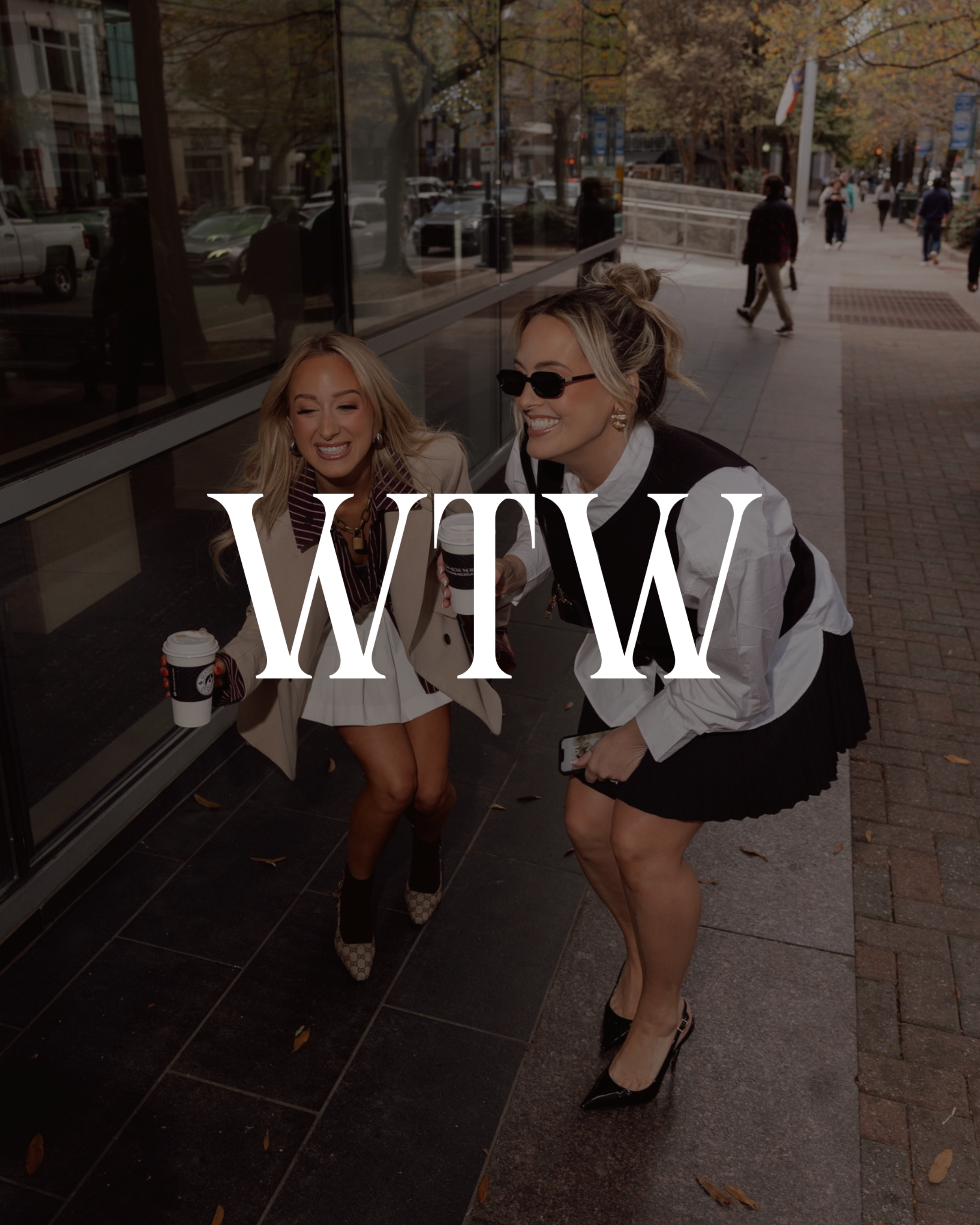 Two women shopping outside on a sidewalk in autumn, holding coffee cups and smiling. One woman wears sunglasses and a white shirt, the other wears a beige blazer and patterned boots.
