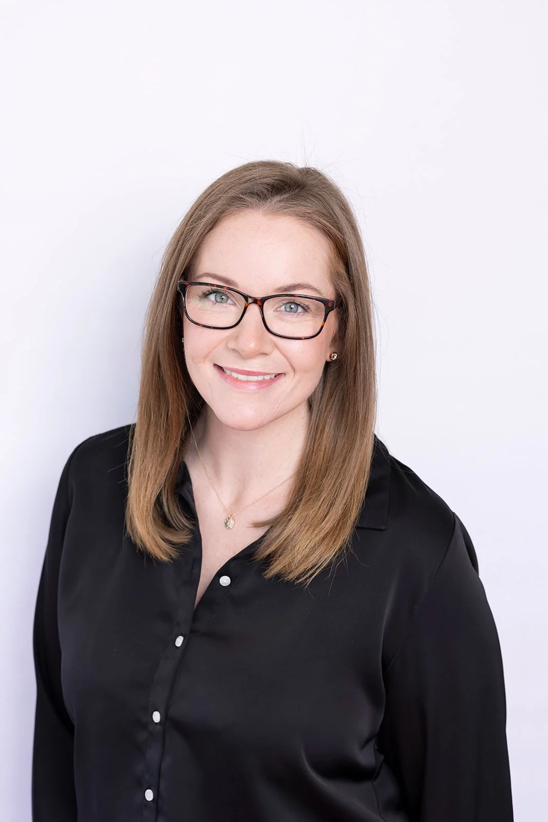A woman with light brown hair, glasses, and a black blouse, smiling against a plain white background.