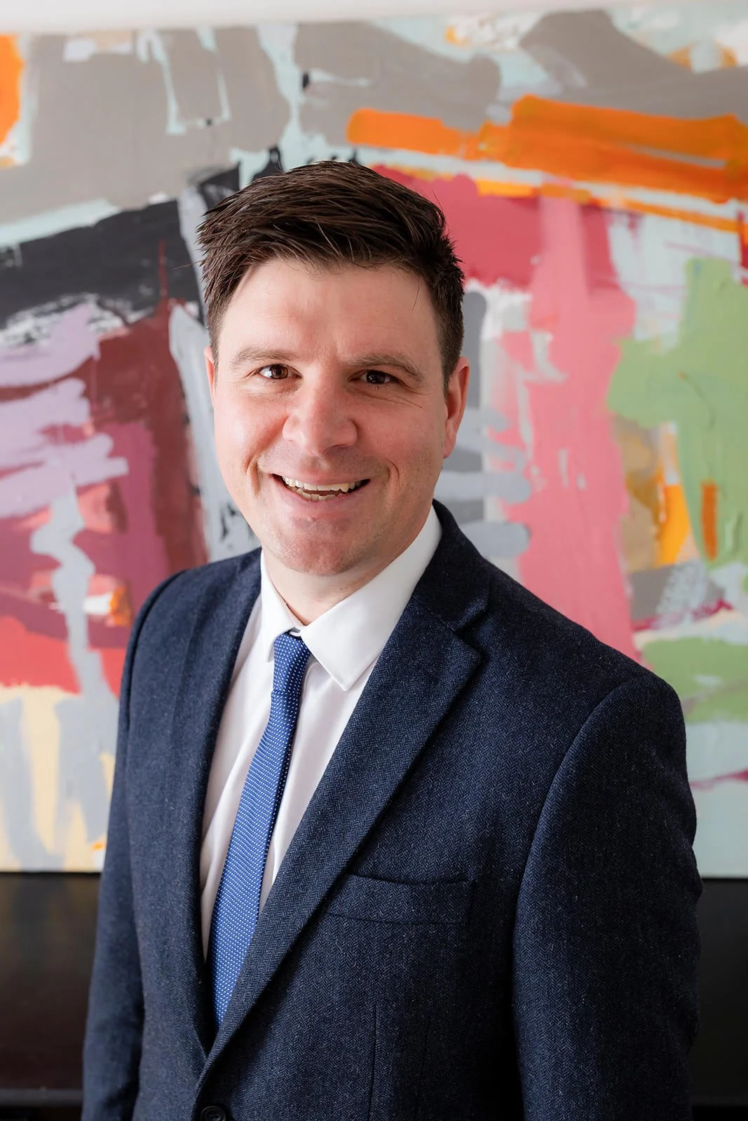 A smiling man in a dark suit, white shirt, and blue tie standing in front of an abstract colorful painting.