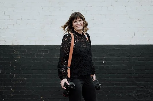 Woman holding two cameras, standing outdoors against a black and white brick wall.
