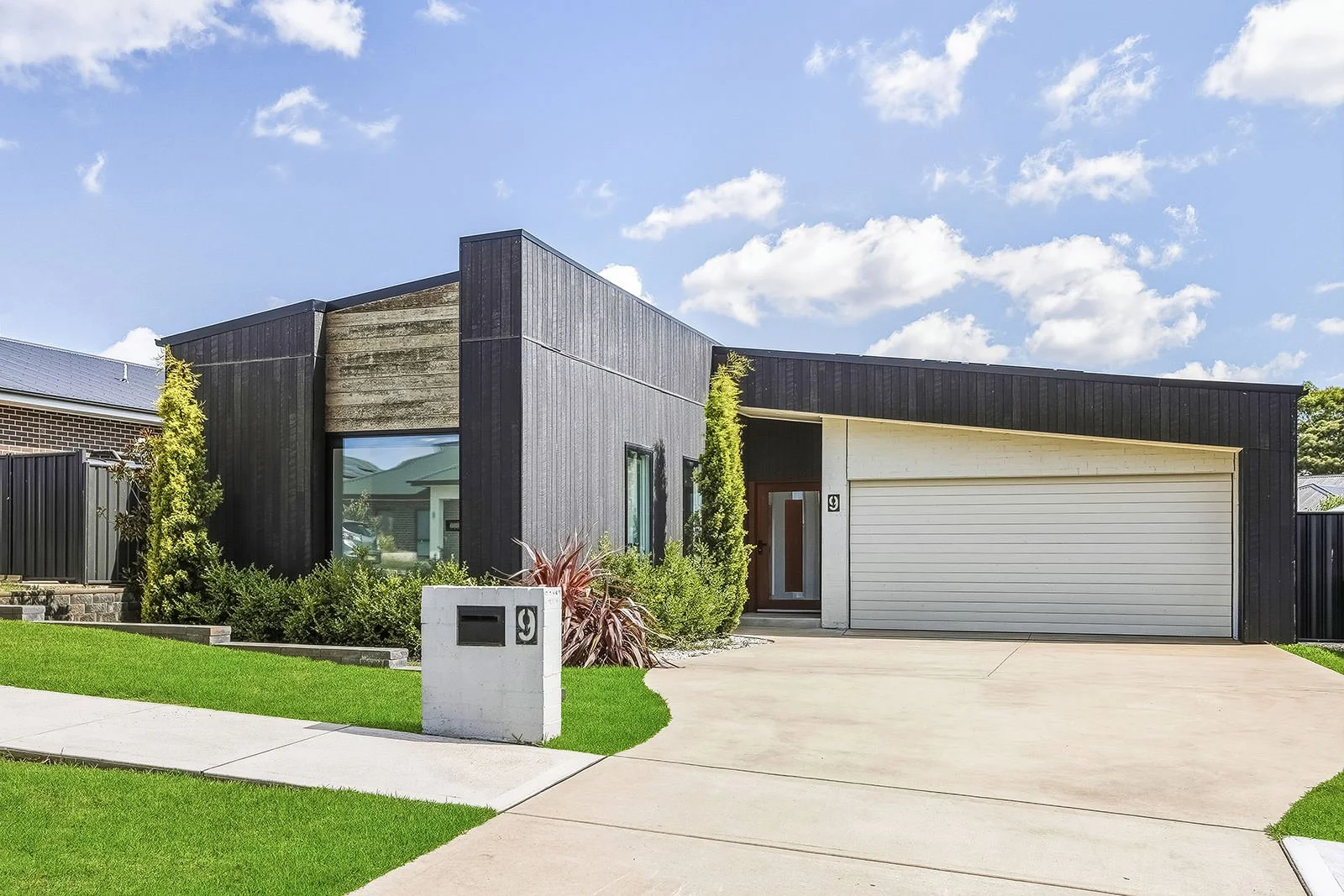 Modern house with black and white exterior, concrete driveway, green lawn, and landscaped plants, under a blue sky with clouds.
