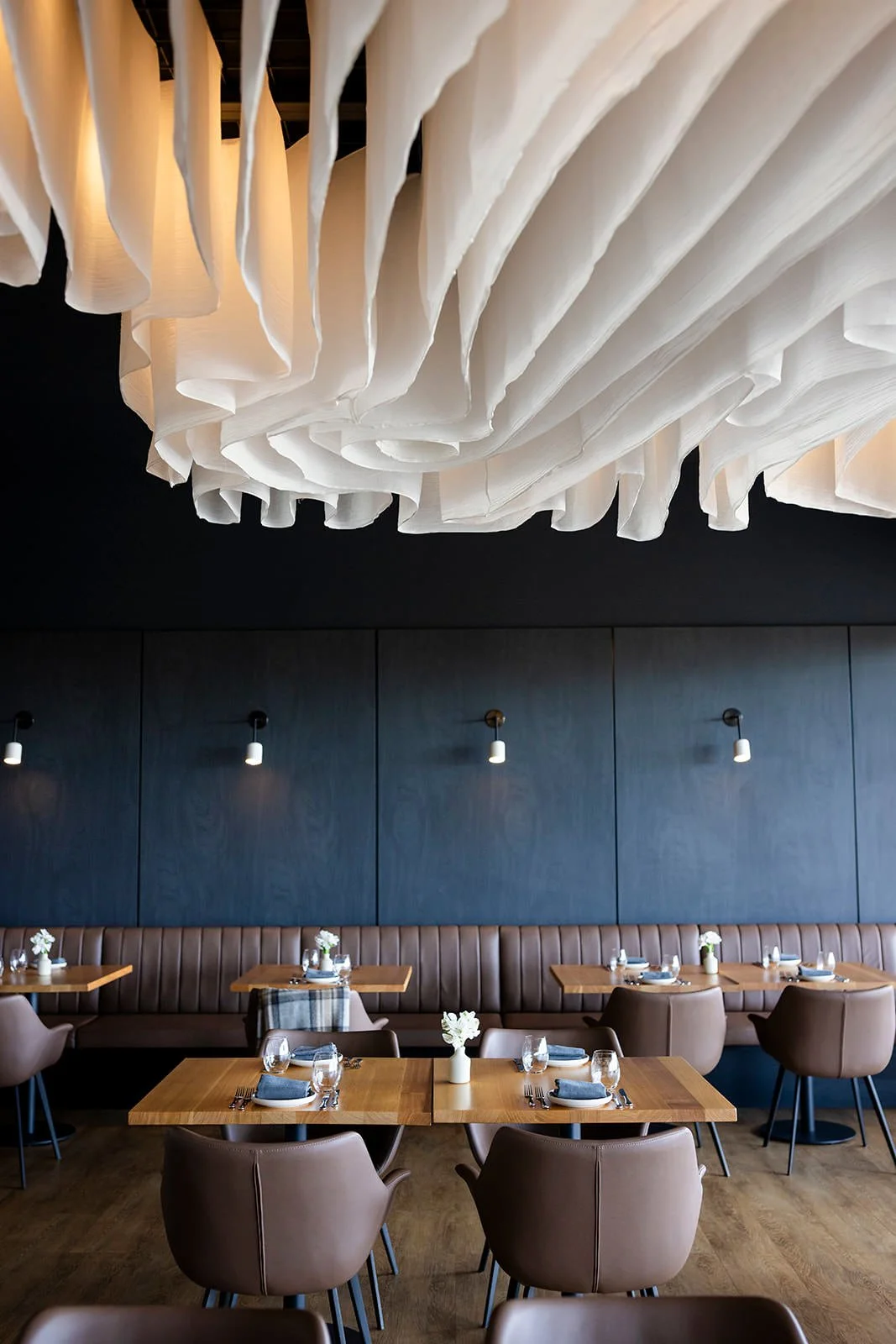 Empty dining area with wooden tables, beige chairs, a brown leather booth, dark blue walls, and a decorative ceiling installation.
