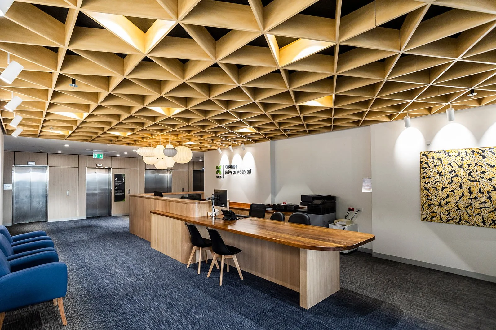 Hospital reception area with a long wooden desk, black chairs, blue armchairs, a colorful artwork on the wall, and a decorative wooden ceiling.