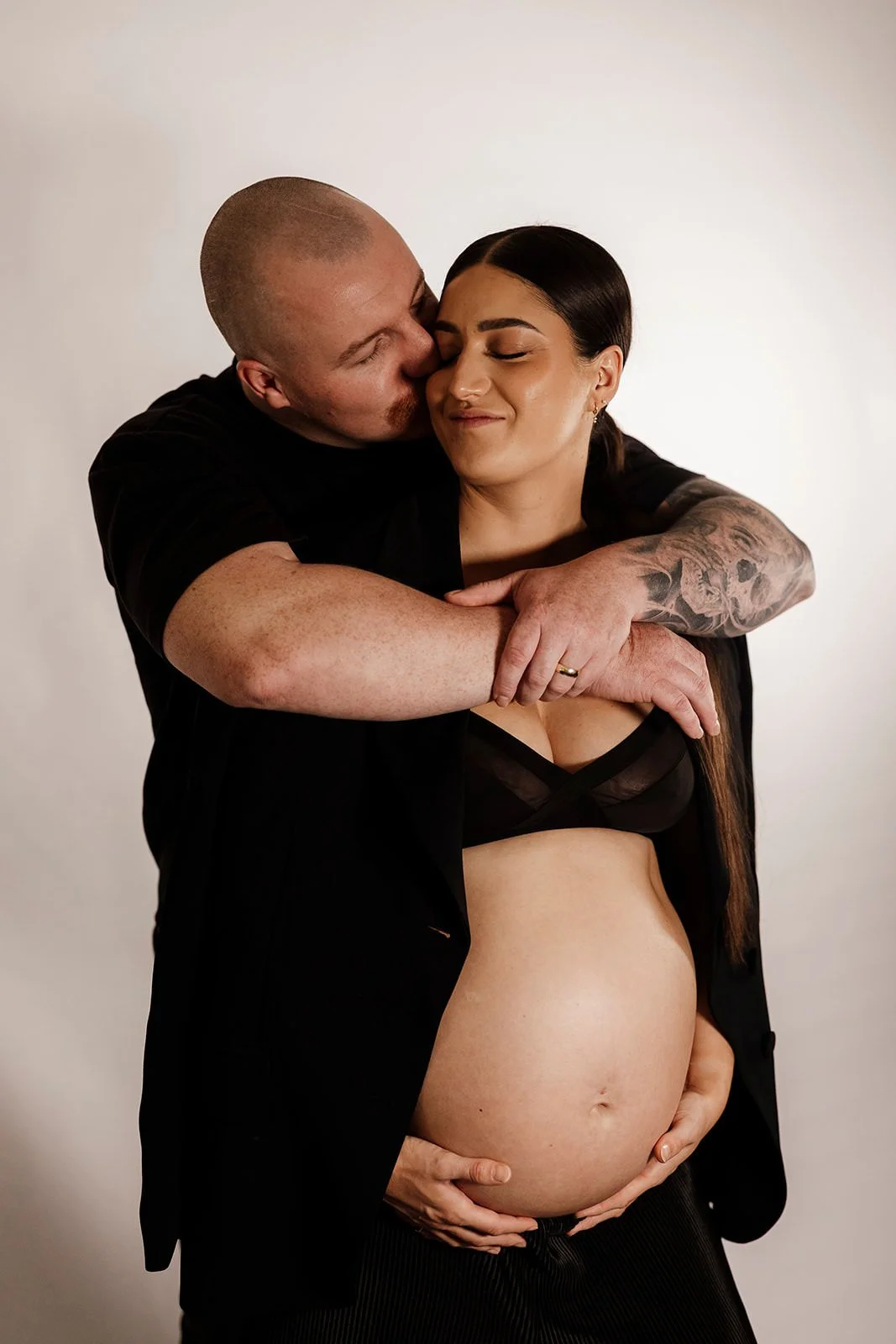 A man and woman embracing, with the woman pregnant and showing her bare belly. The man is kissing her cheek with eyes closed, and the woman has a content smile. They are standing against a plain, light-colored background.