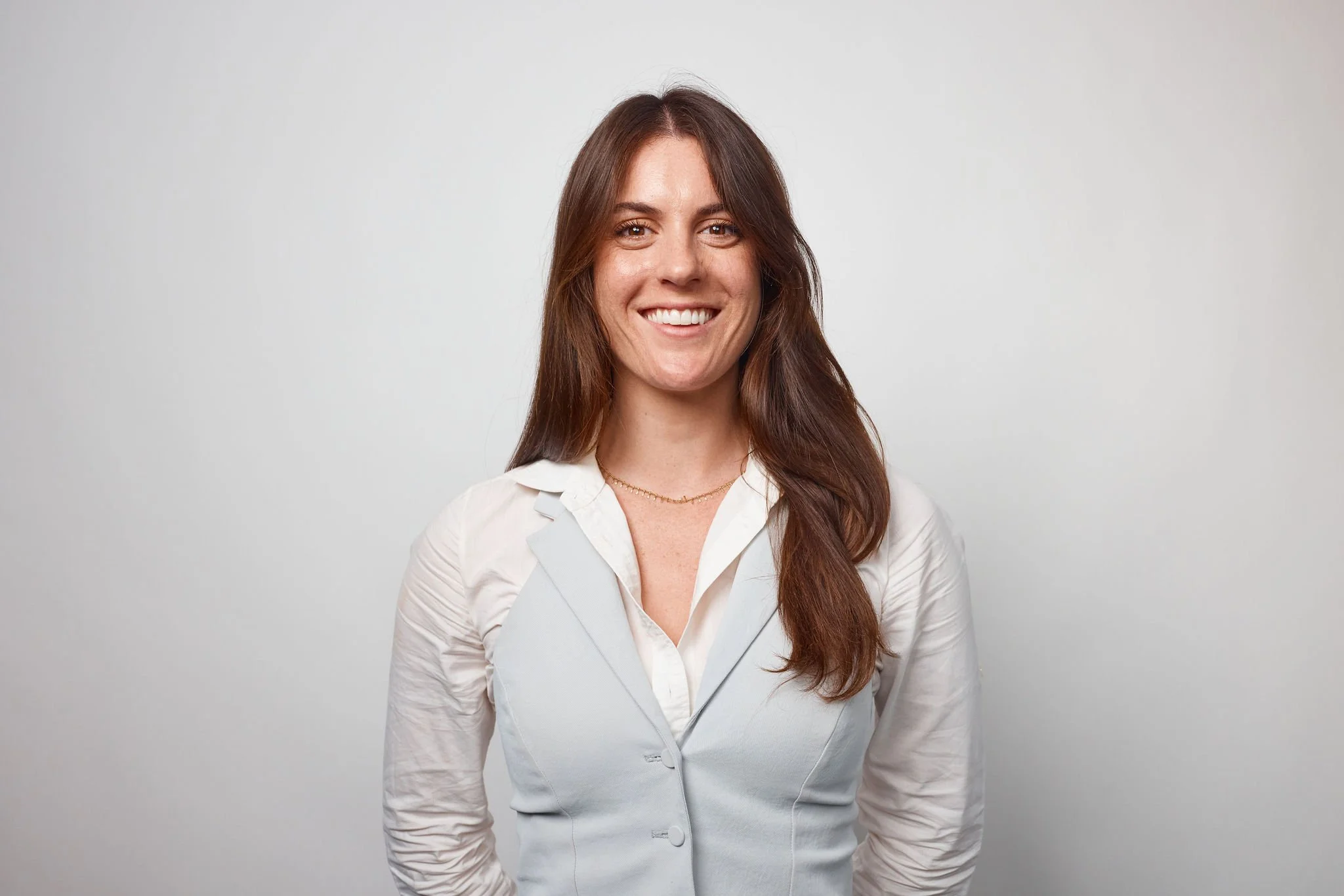 A portrait of a smiling woman with long brown hair, wearing a white blazer and a gold necklace, standing against a plain light gray background.