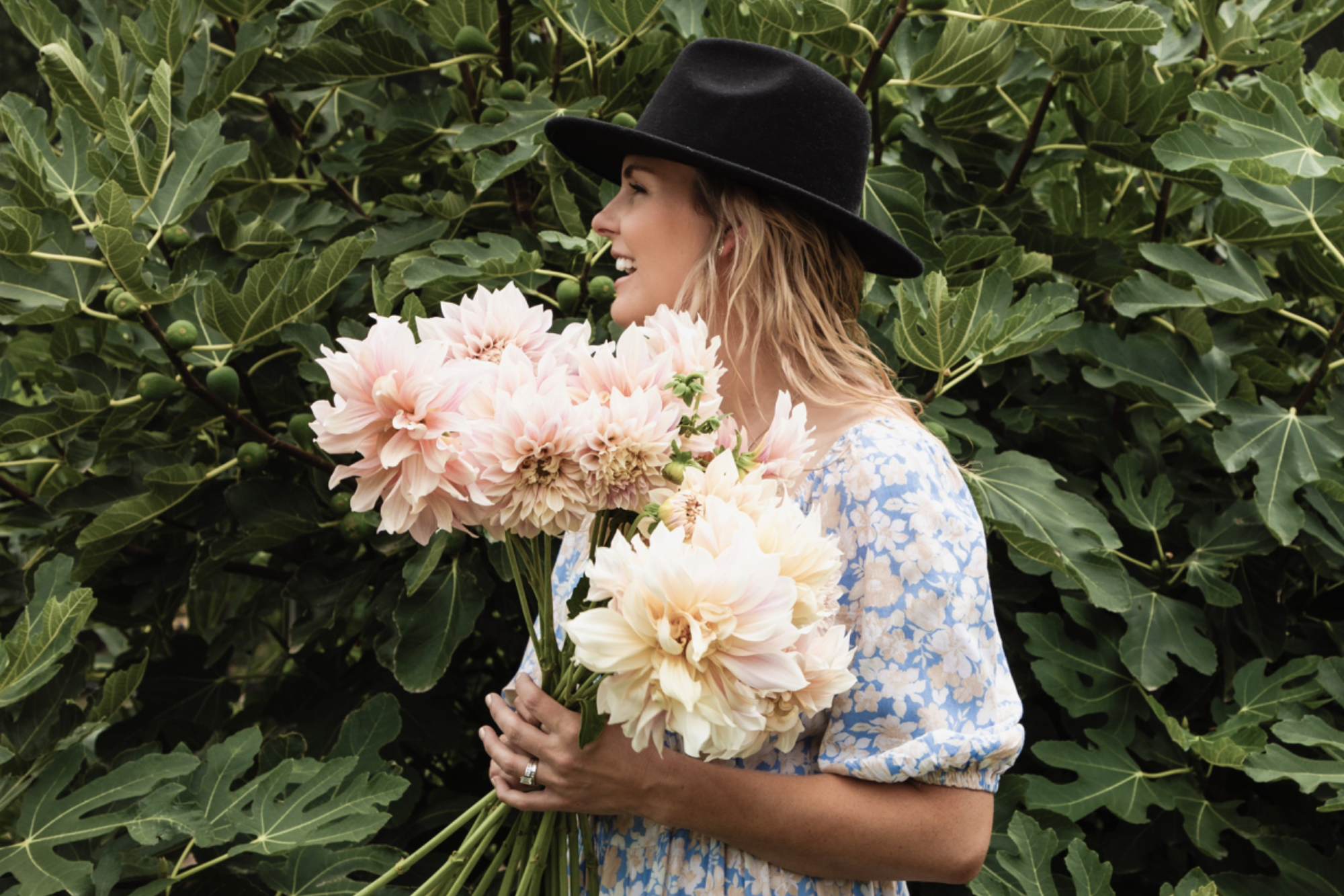 Woman in floral dress holding bouquet of dahlias, wearing black hat, surrounded by green foliage.
