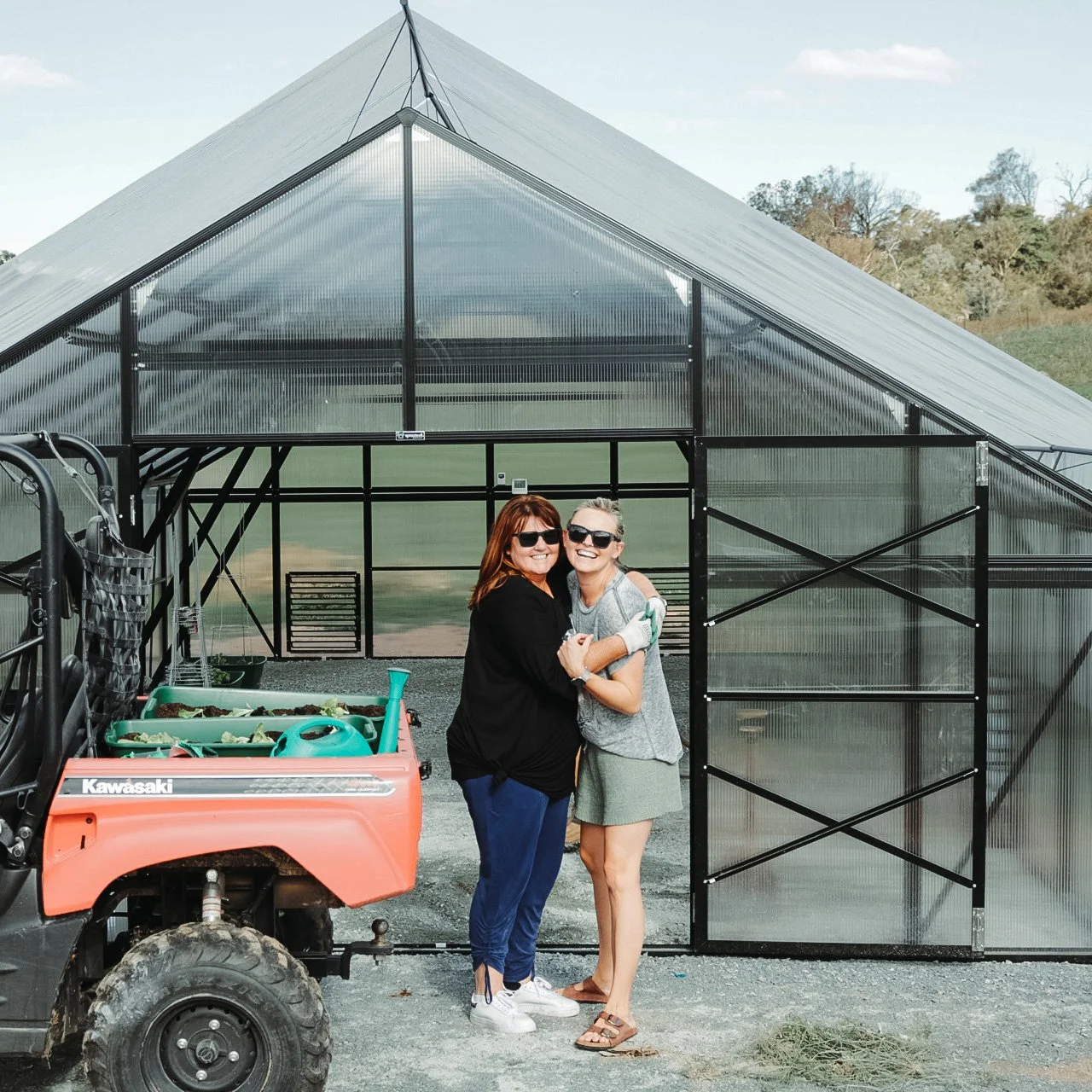 Two people smiling and hugging in front of a greenhouse with a utility vehicle nearby, on a sunny day.