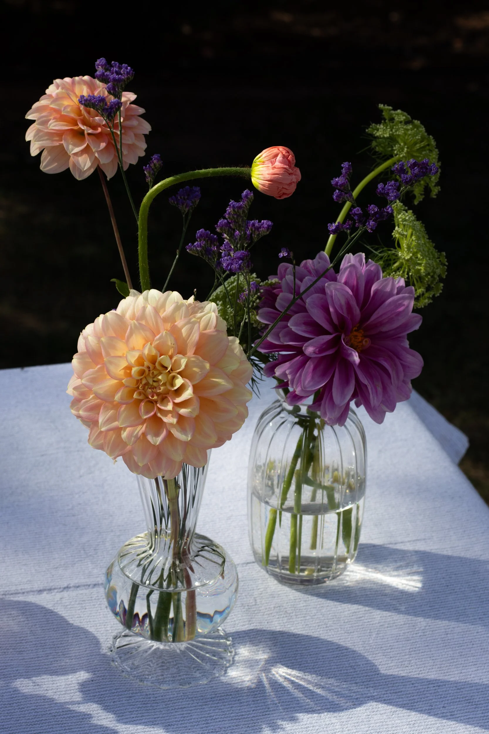 Two vases on a table with peach and purple dahlias, purple flowers, and greenery against a dark background.