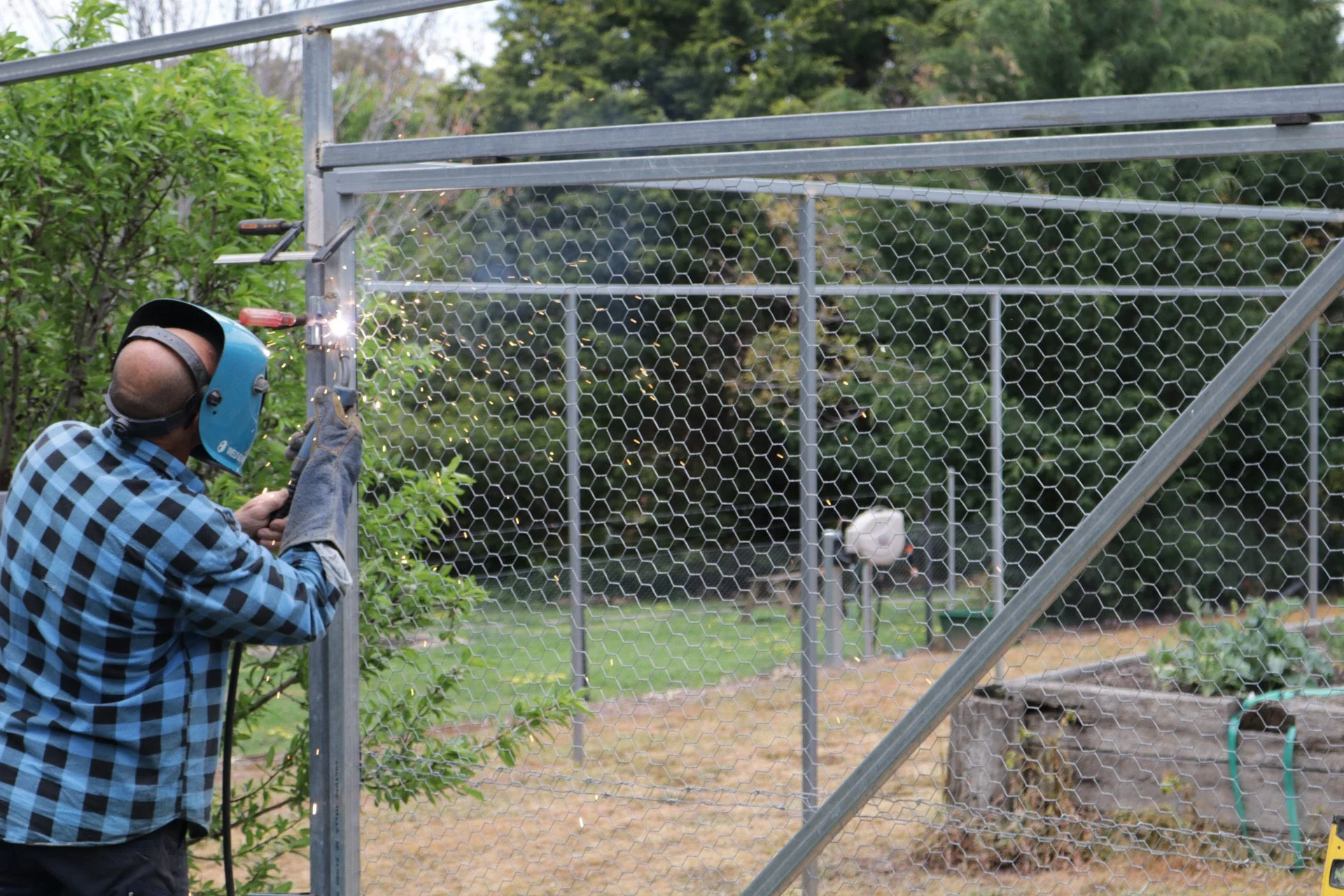 Person welding a metal frame of a fence with hexagonal wire mesh in a garden setting.