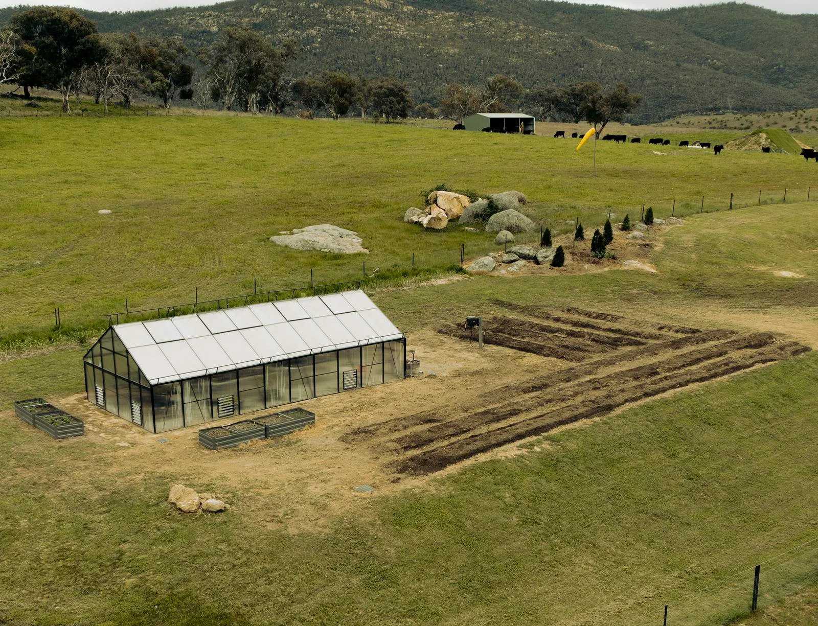 Aerial view of a greenhouse on a rural field with surrounding garden beds and distant hills.