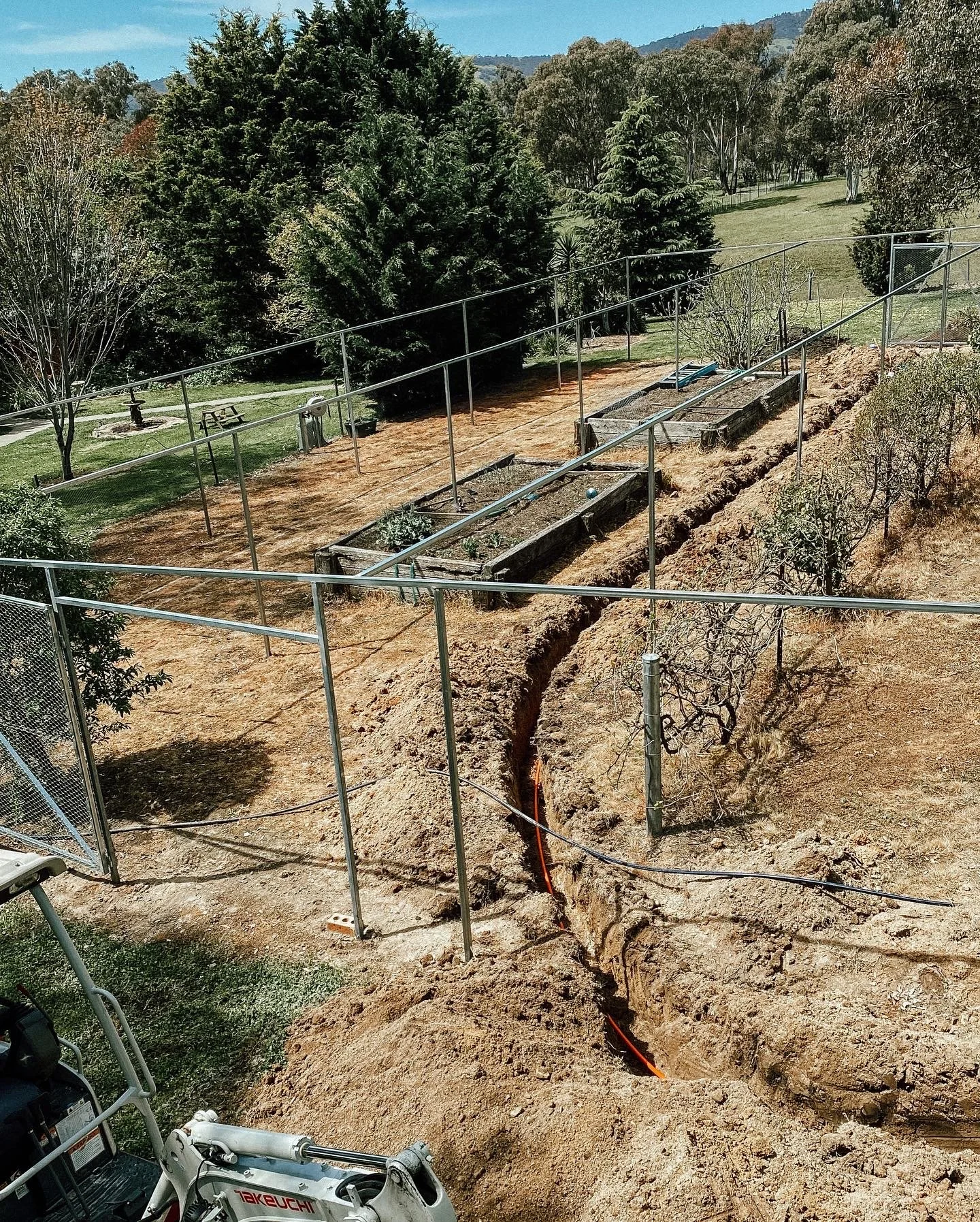Garden area with fencing and a trench under construction, surrounded by trees and grassy landscape.