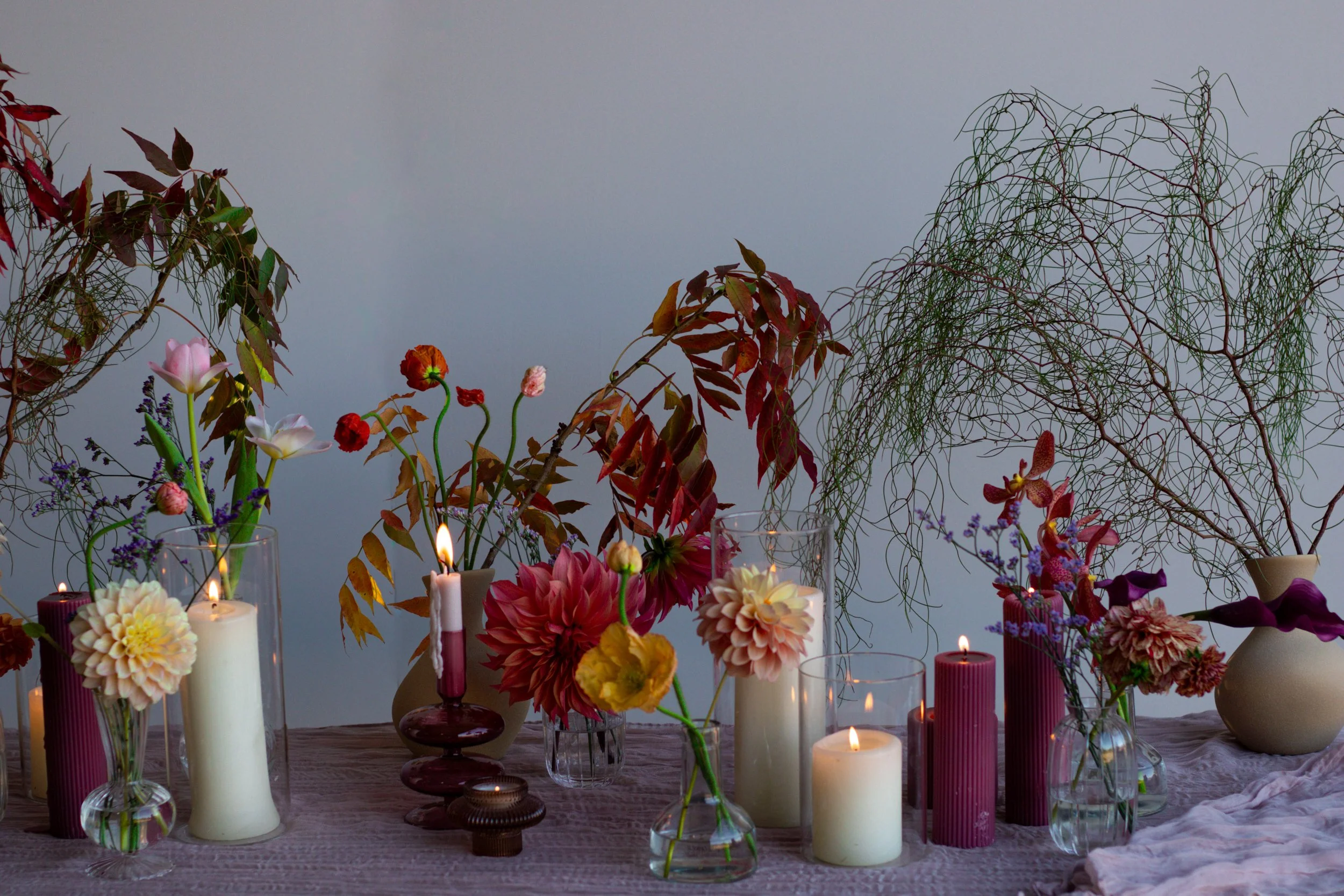 A table display with various colorful flowers, including dahlias and orchids, arranged in glass vases. Multiple candles are lit, distributed throughout the arrangement, creating ambient lighting. The background includes leafy plants.