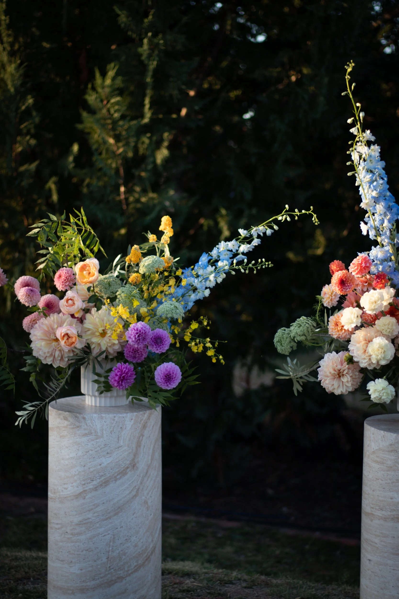 Artistic floral arrangements on marble pedestals outdoors, featuring pink, purple, yellow, and blue flowers with greenery.