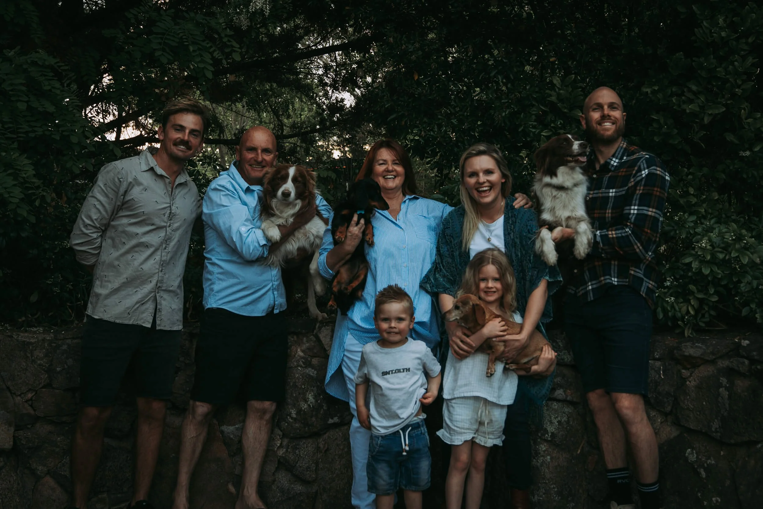 A family posing outdoors with three dogs, surrounded by greenery.