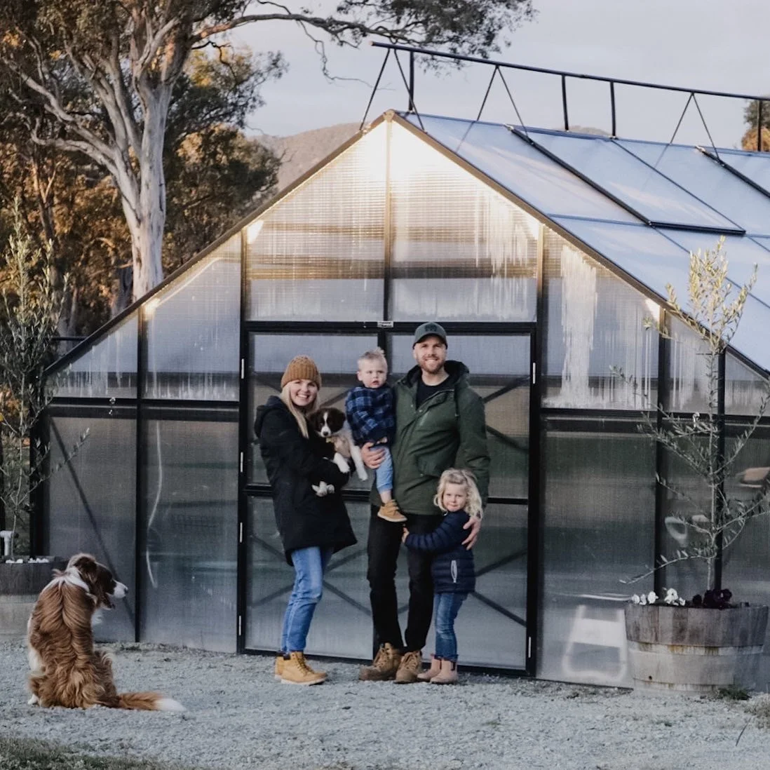 A family with two children and a dog standing in front of a greenhouse.