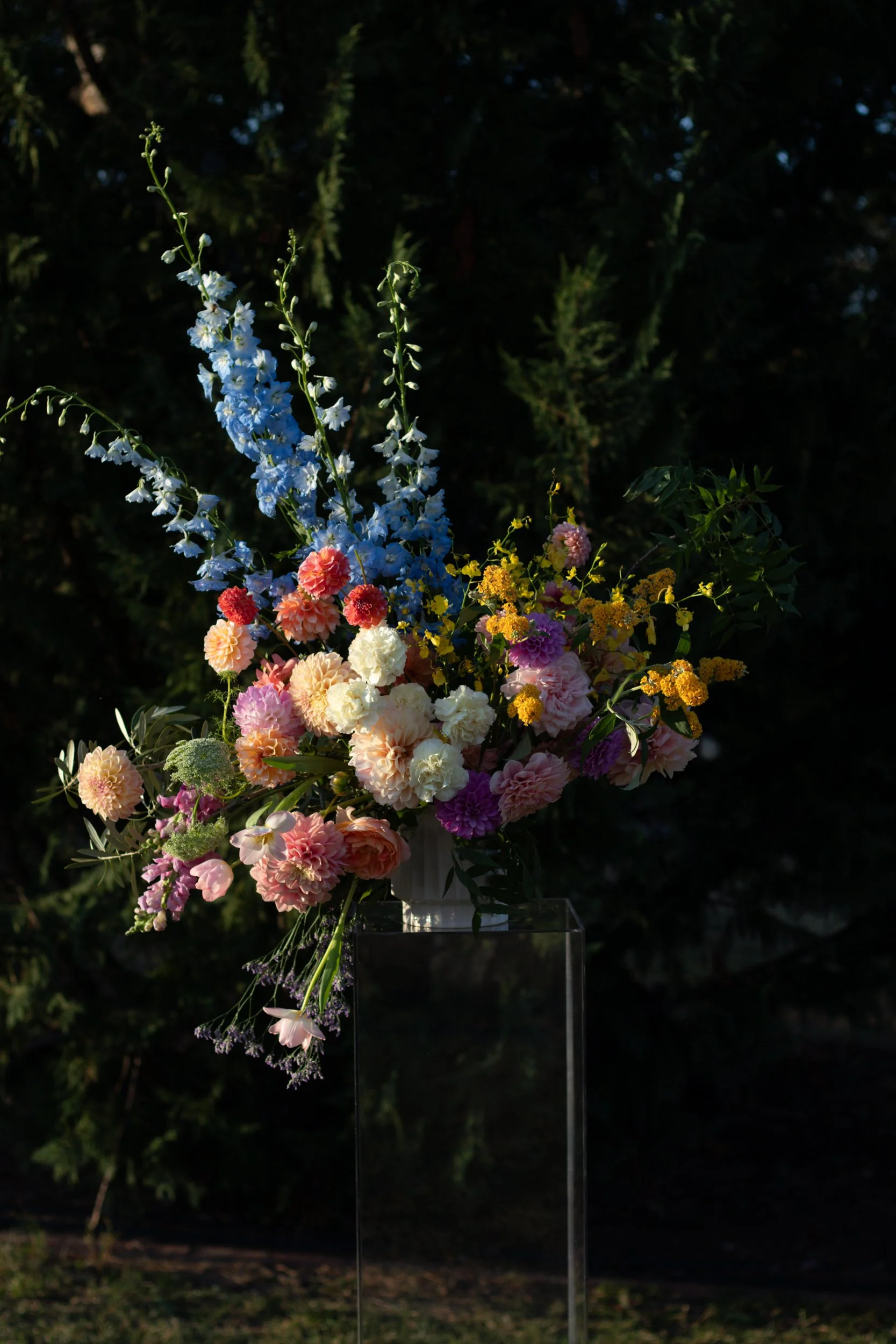 Colorful floral arrangement with blue delphiniums, pink, peach, and yellow flowers, in white vase, dark green background.