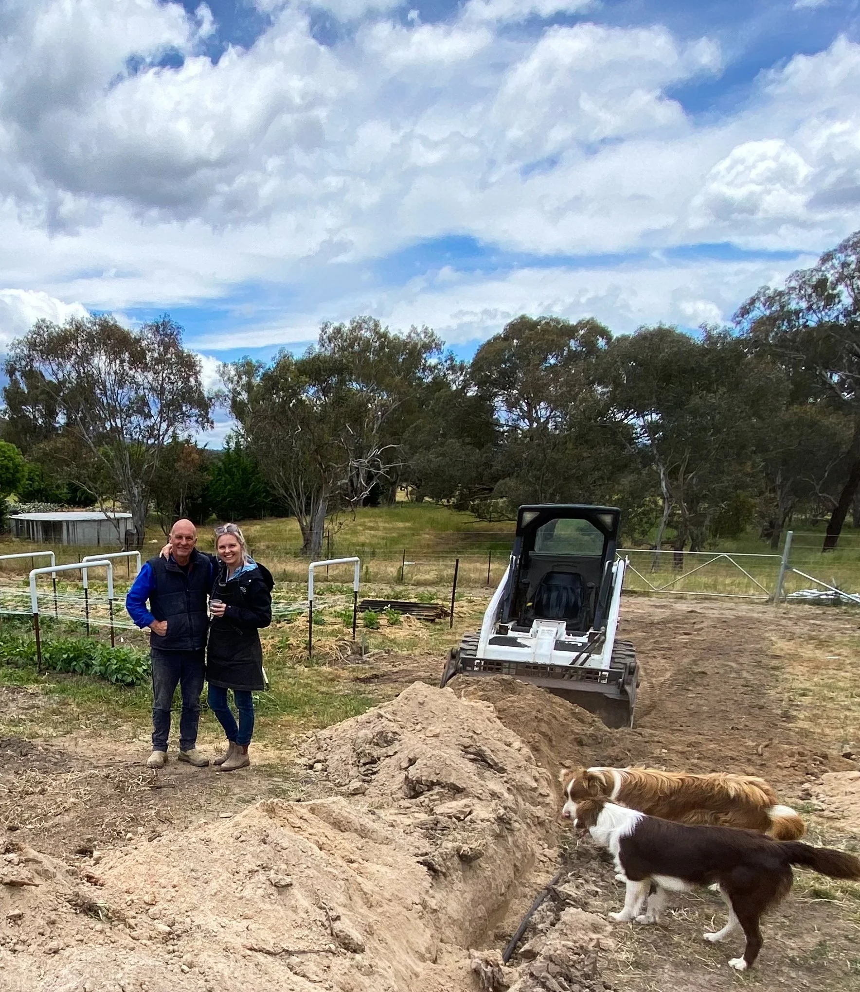 A man and a woman standing together near a dug trench with two dogs nearby and a small excavator in the background in a rural setting.