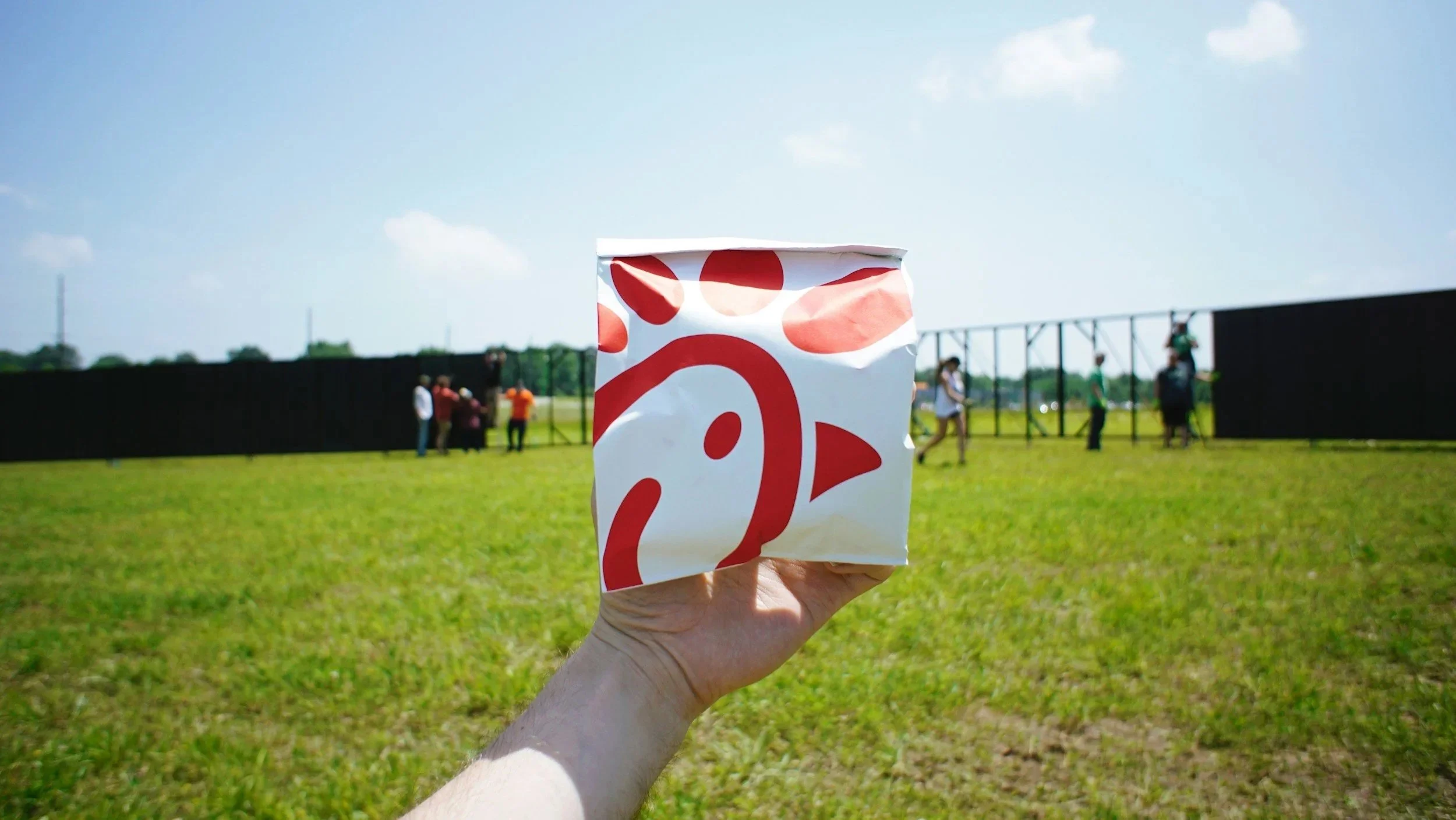 Hand holding a Chick-fil-A cup at an outdoor event on a sunny grassy field