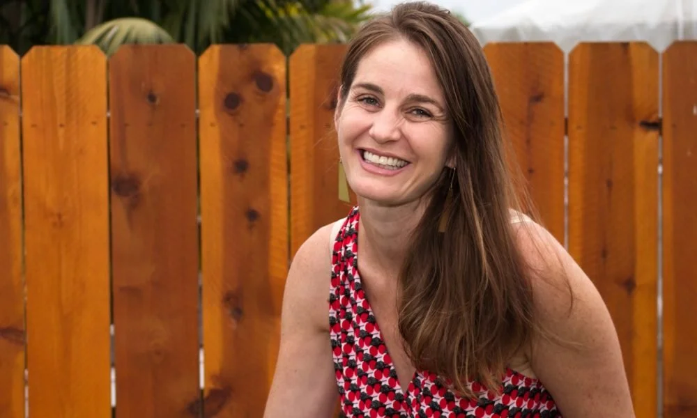 Owner Amanda Manger, a woman smiling with long brown hair smiling outdoors in front of a wooden fence, wearing a sleeveless top with red, black, and white patterns.