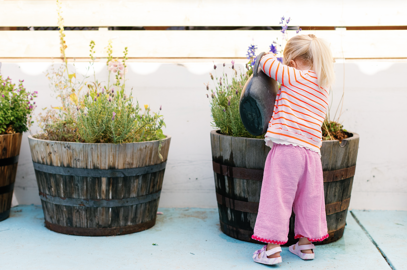 A young girl with blonde hair watering plants in large wooden planters on a patio.