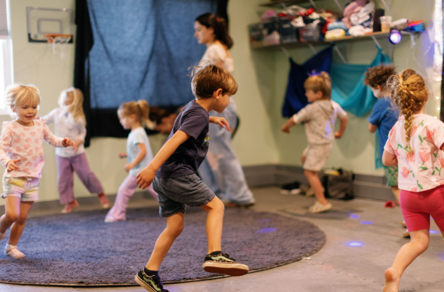 Young children dancing and playing in preschool classroom for movement class, with a woman supervising in the background, and various colorful decorations on the wall.