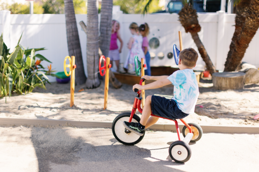A preschooler riding a tricycle in a sandbox area, with a group of three children playing near by and trees in the background.