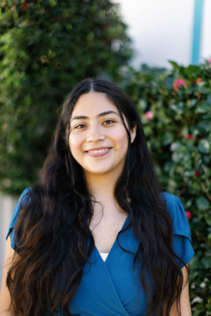 Smiling young woman with long dark hair in a blue top outdoors, greenery in background.