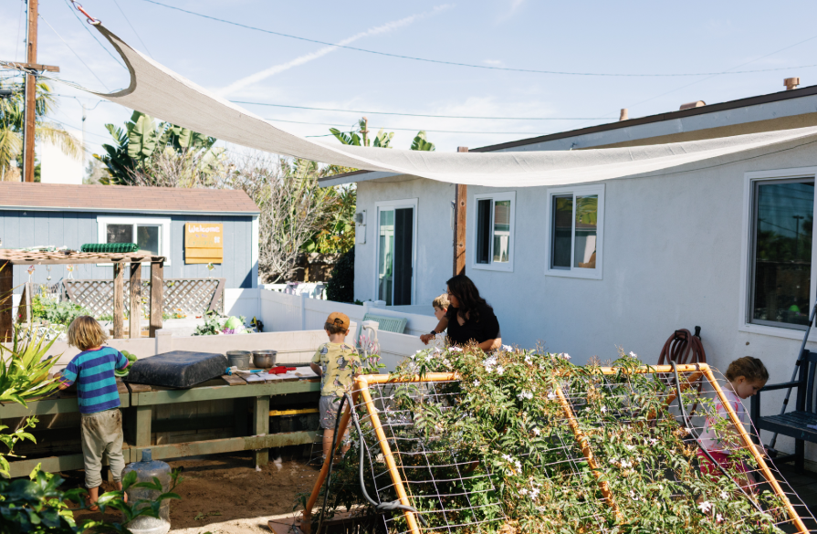 A backyard scene with children and an adult gardening and playing in a sunny outdoor space with plants, a raised garden bed, and a preschool building.