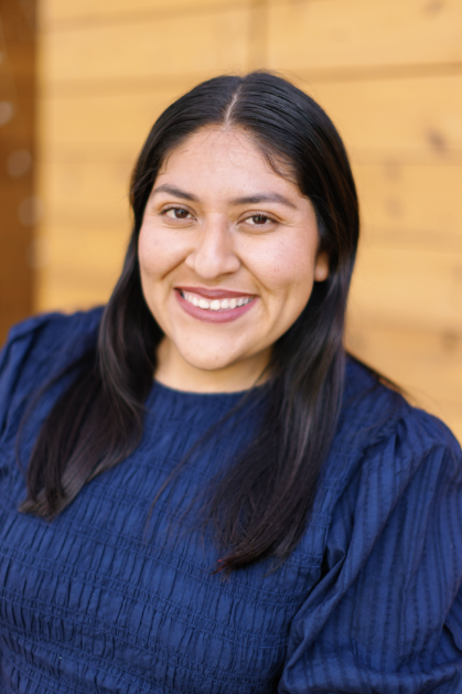 A woman with long dark hair wearing a blue top smiling in front of a wooden wall.